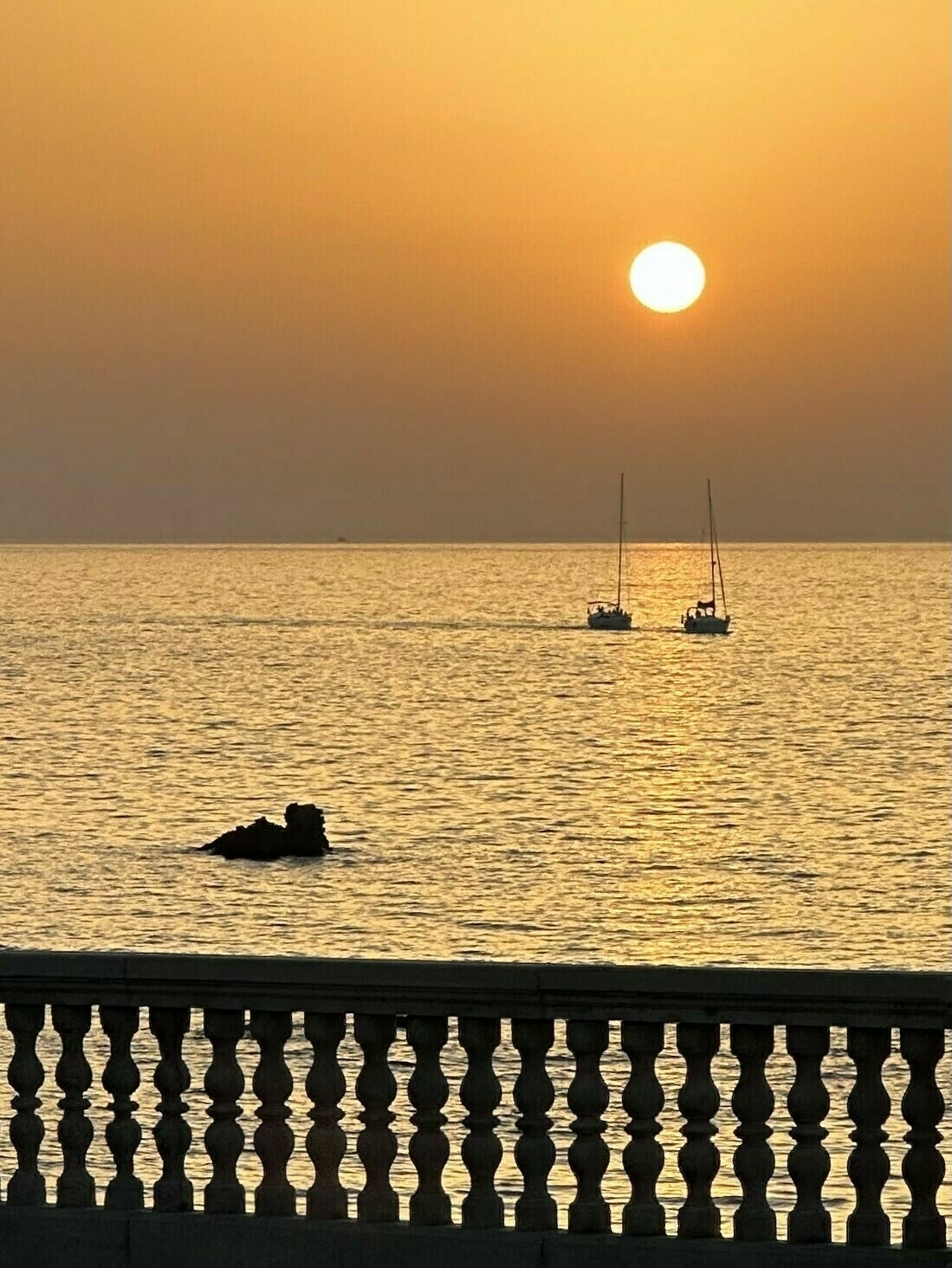 Sunset over the sea, two sailing boats in the distance under the glowing sun, a railing silhouetted in the foregrounds 