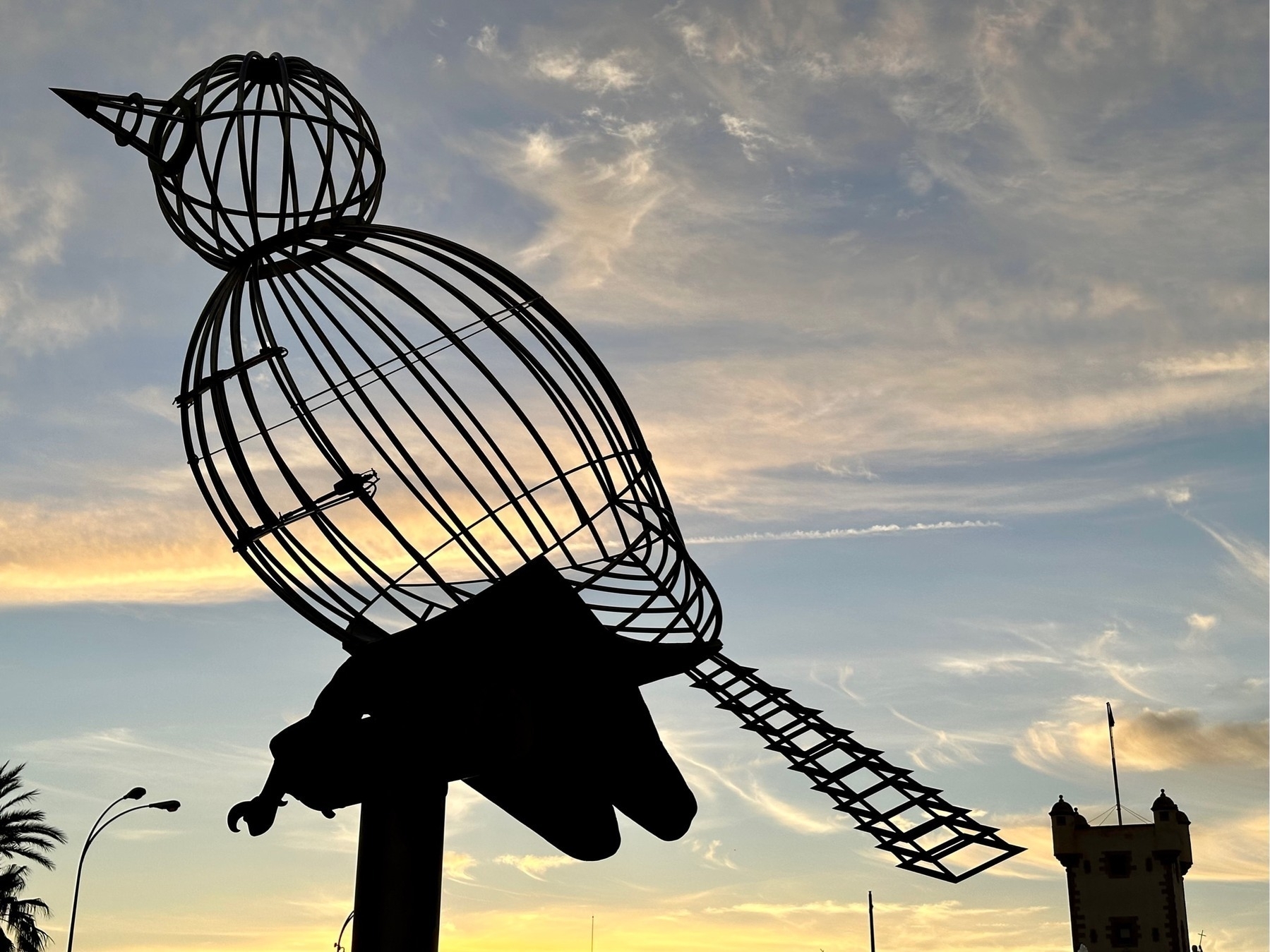 Wireframe bird sculpture in silhouette against a setting sky, a turret in the background