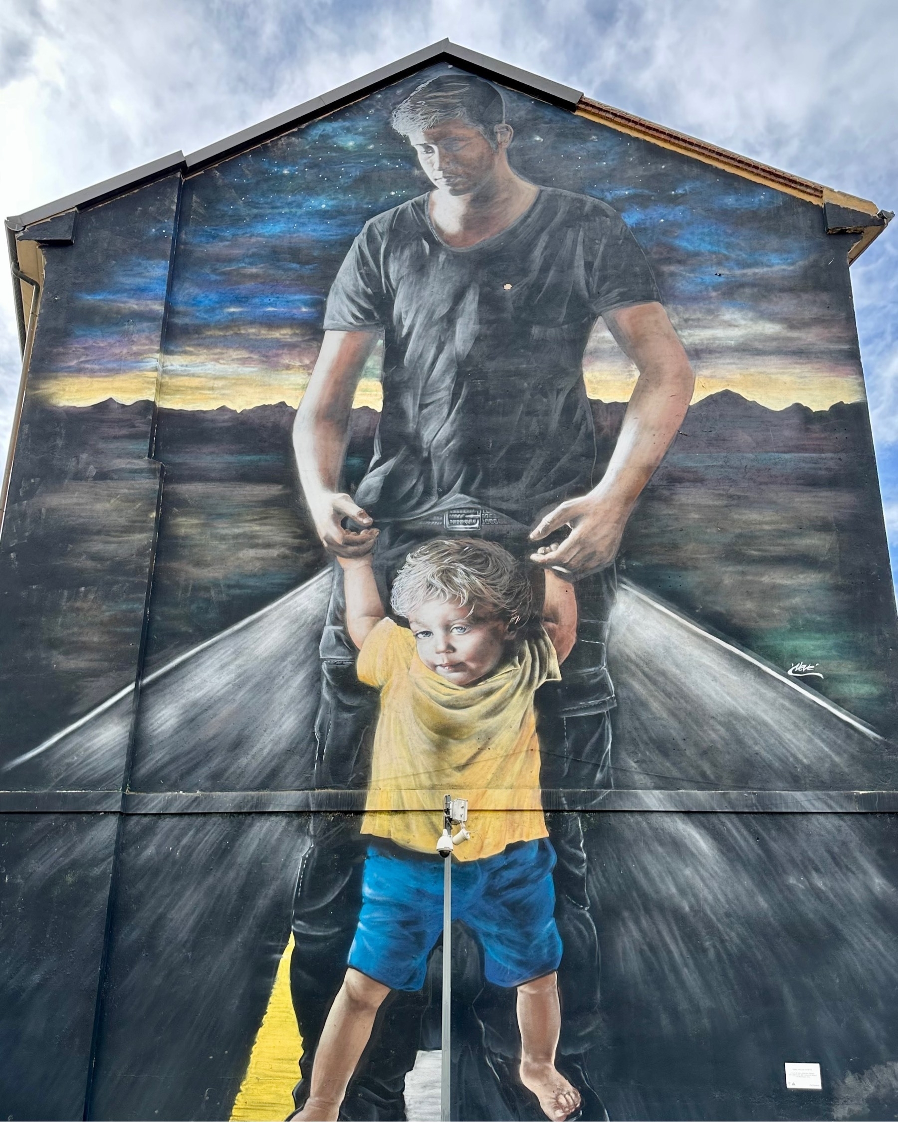 Lifelike mural on the side of a building, showing a man leading a child down a road