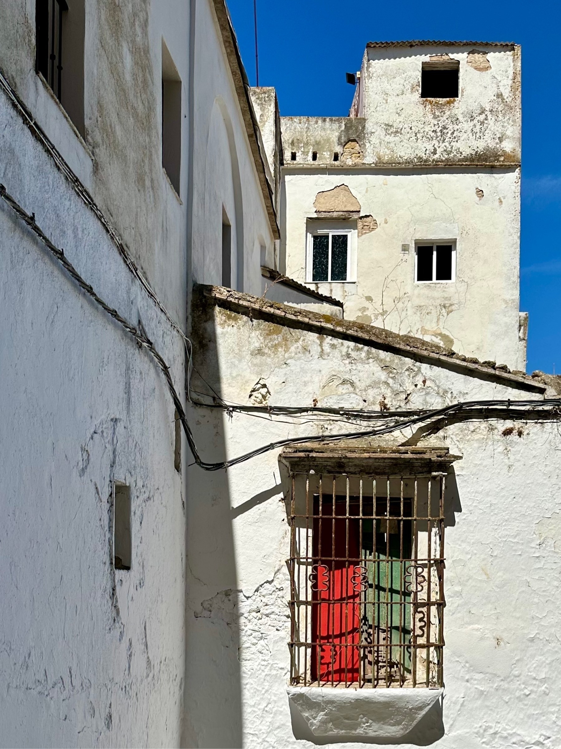 White 17th-century Hispanic buildings close up against a blue sky, one main window covered up with two wooden boards; one red on the left, and one green on the right.