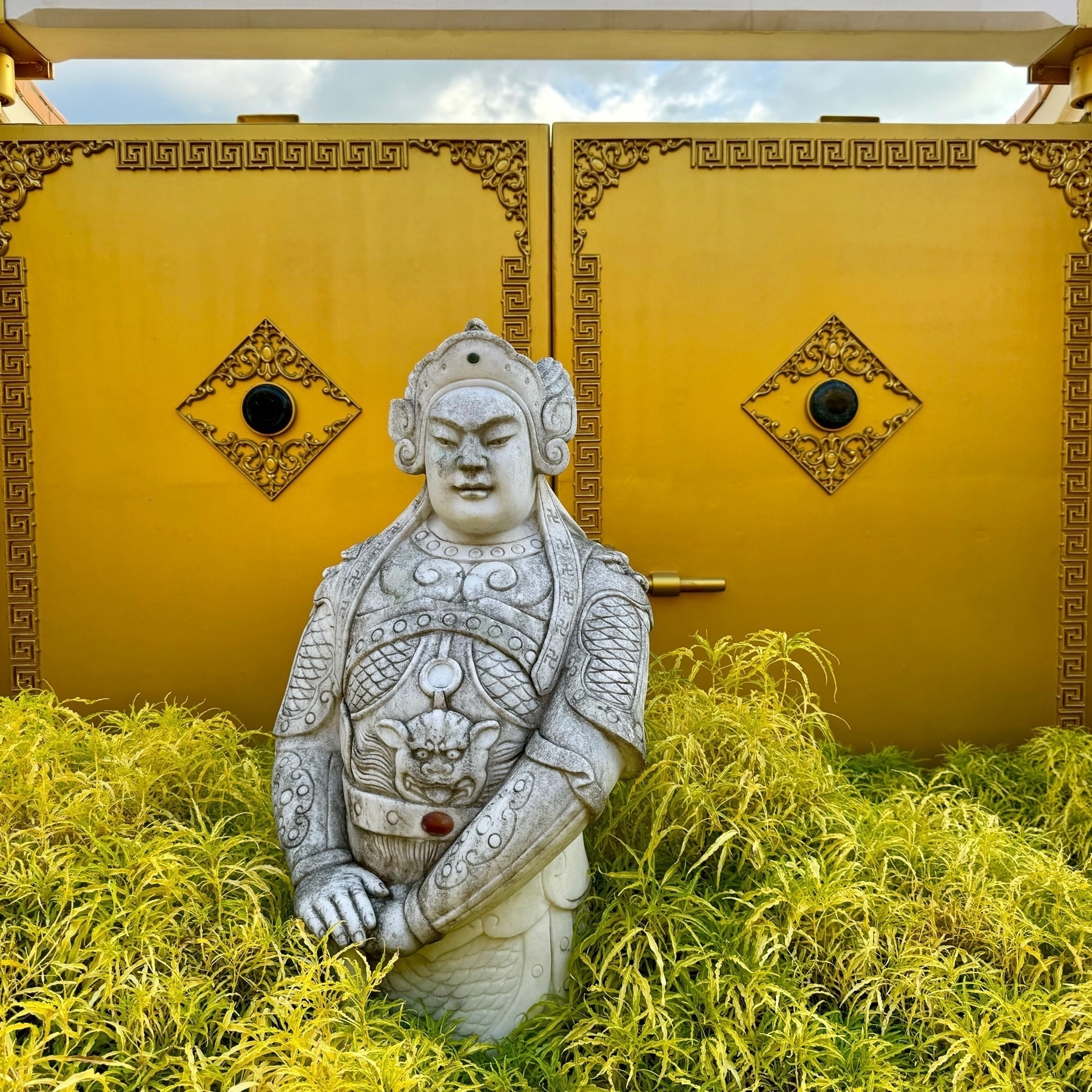 A religious statue in a temple, standing waist-down in a light-green bush, with a backdrop of two yellow decorated doors