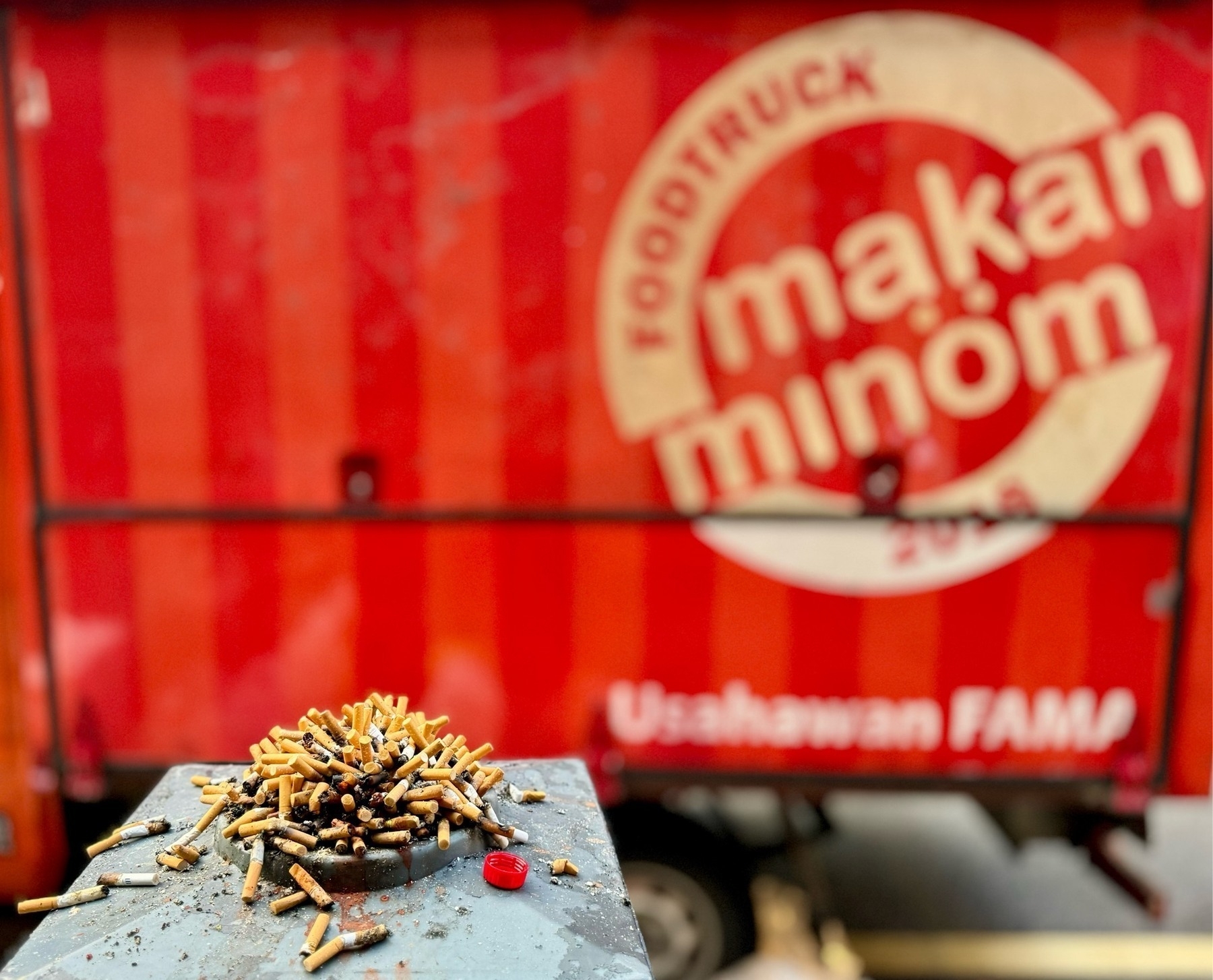 A huge pile of cigarette butts on a disposal stand, in front of the side of a food truck, in red with white writing