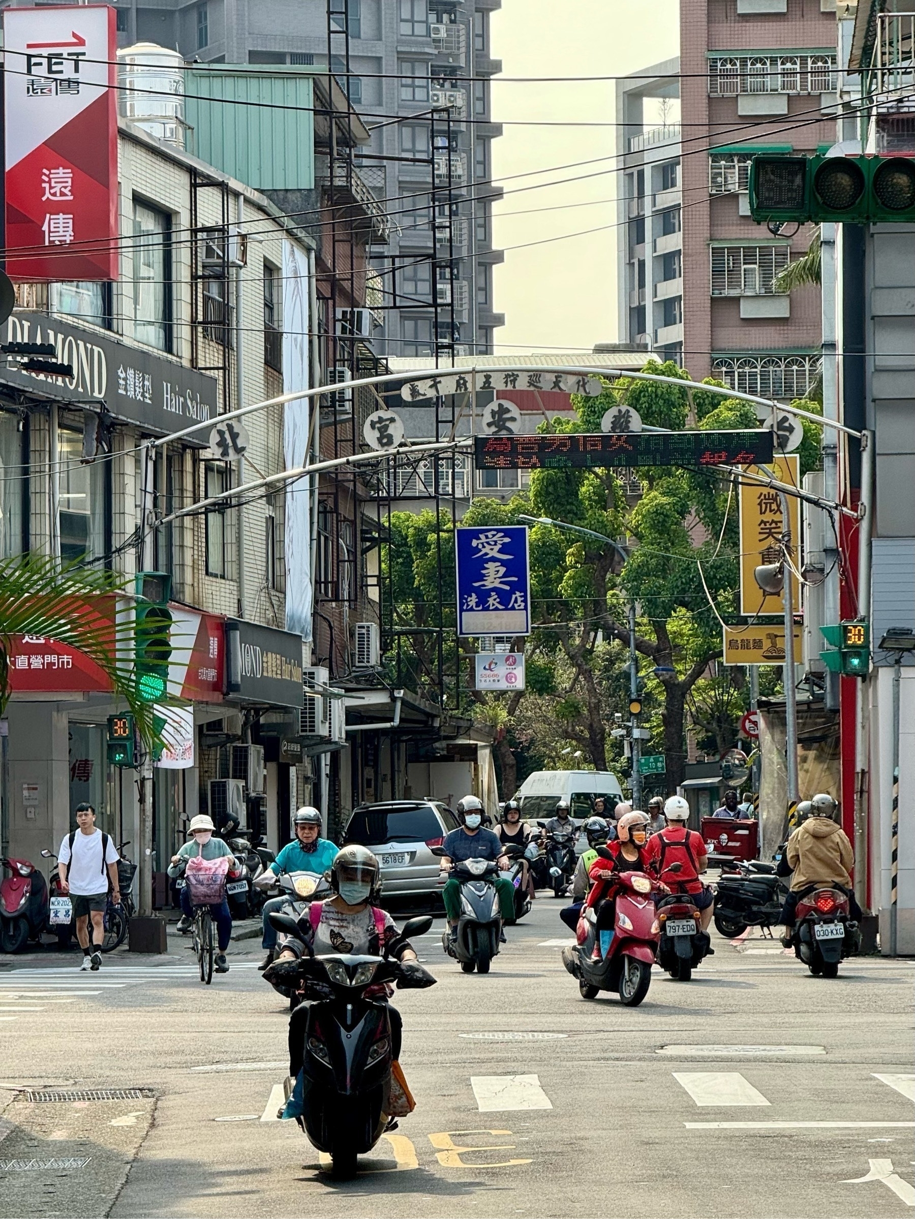 A small road junction, with several motorcycles coming towards the camera, and a blue sign hanging in the distance reading 愛妻洗衣店