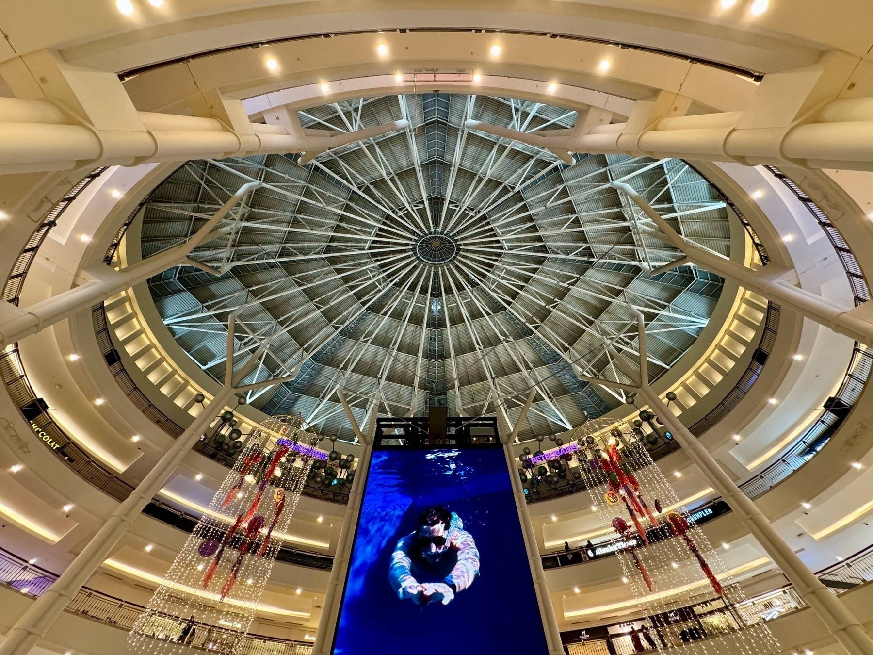 Looking up towards the central ceiling dome in a shopping mall, with a large screen in front. On the screen a man is shown snorkelling towards us through blue water