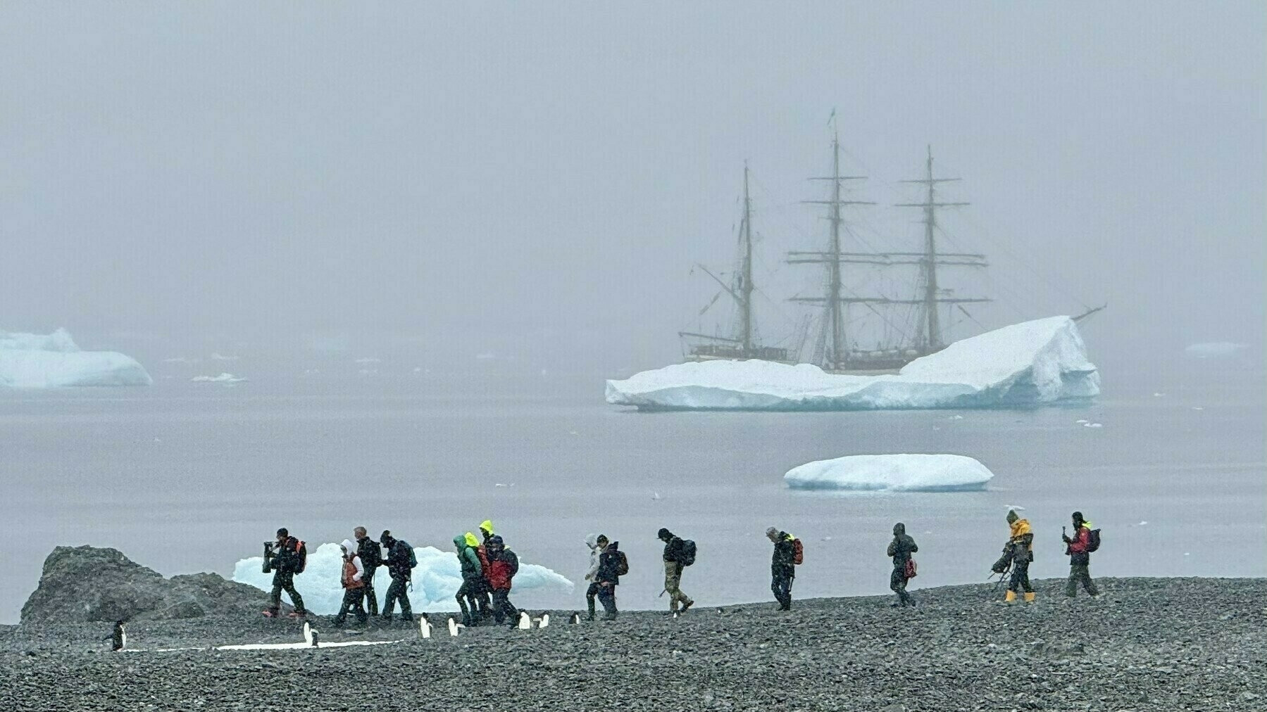 Dutch tall (sailing) ship behind an iceberg in the background, with several people and penguins walking along the beach in the snow in front