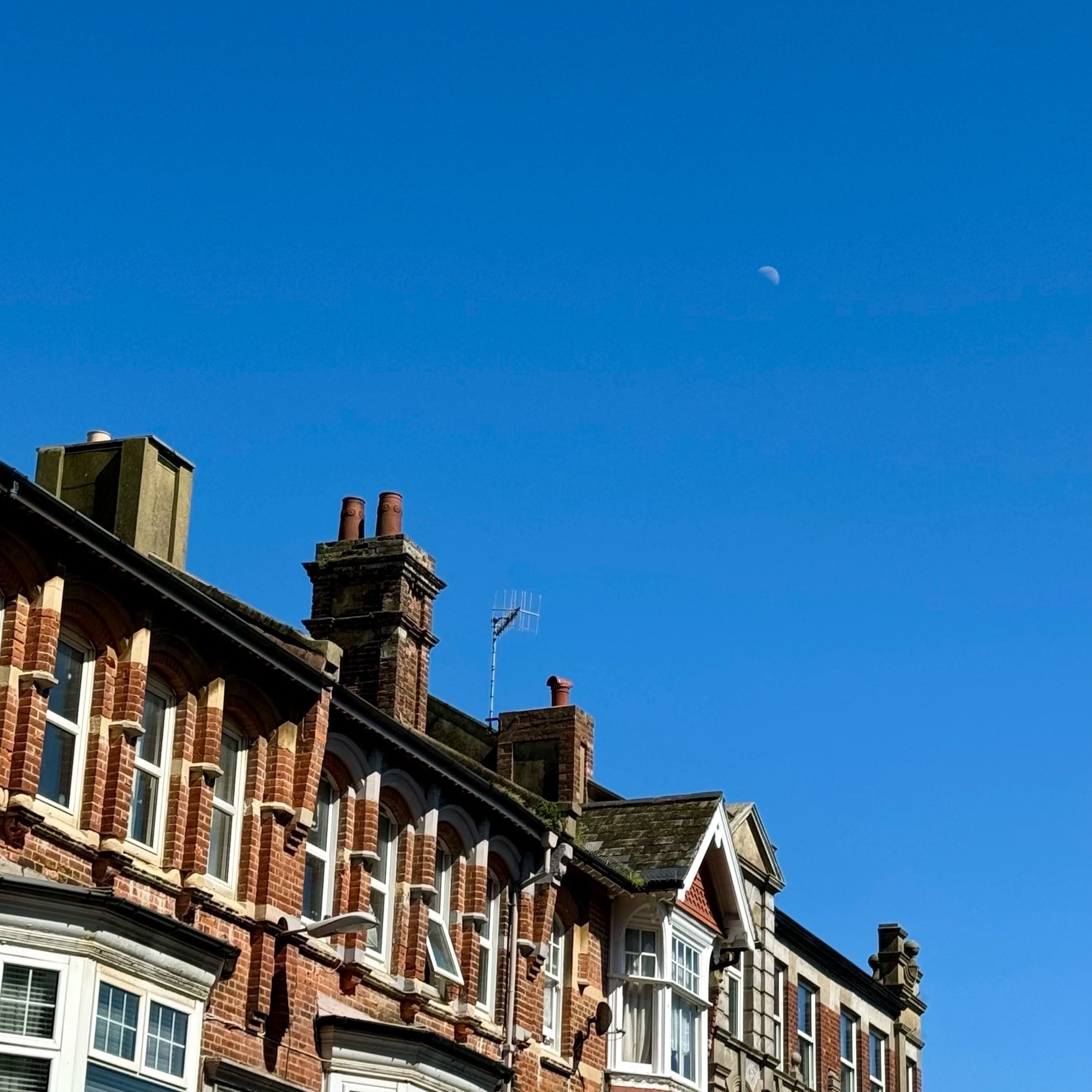 The slight "smiling" moon during the daytime, in a deep blue sky over brick townhouses