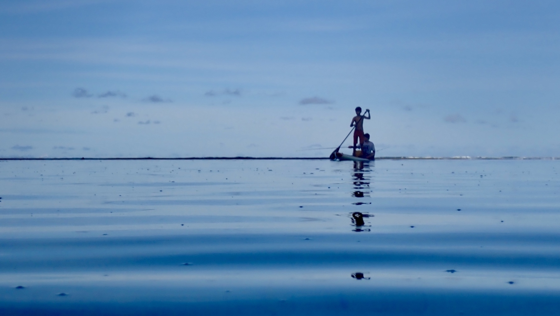 Blue sea, blue sky; horizon through the middle. Two boys on an SUP board; one stood up and paddling, one sat down and fishing