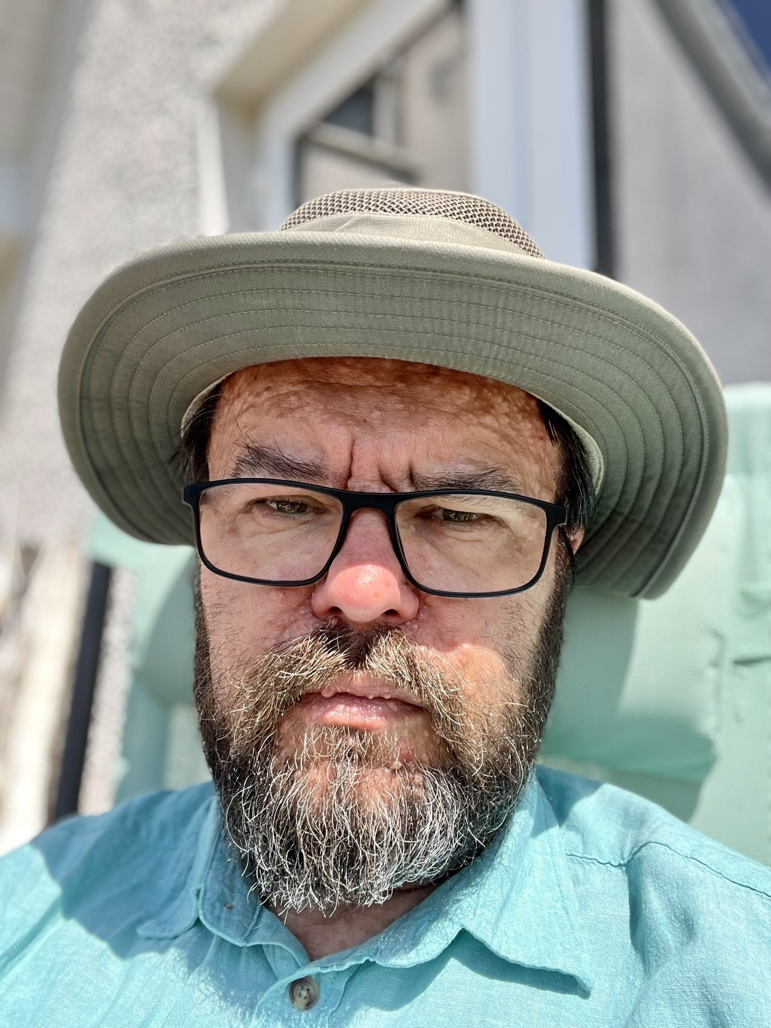 A grumpy bearded old man looks morosely at the camera, a hat atop his head