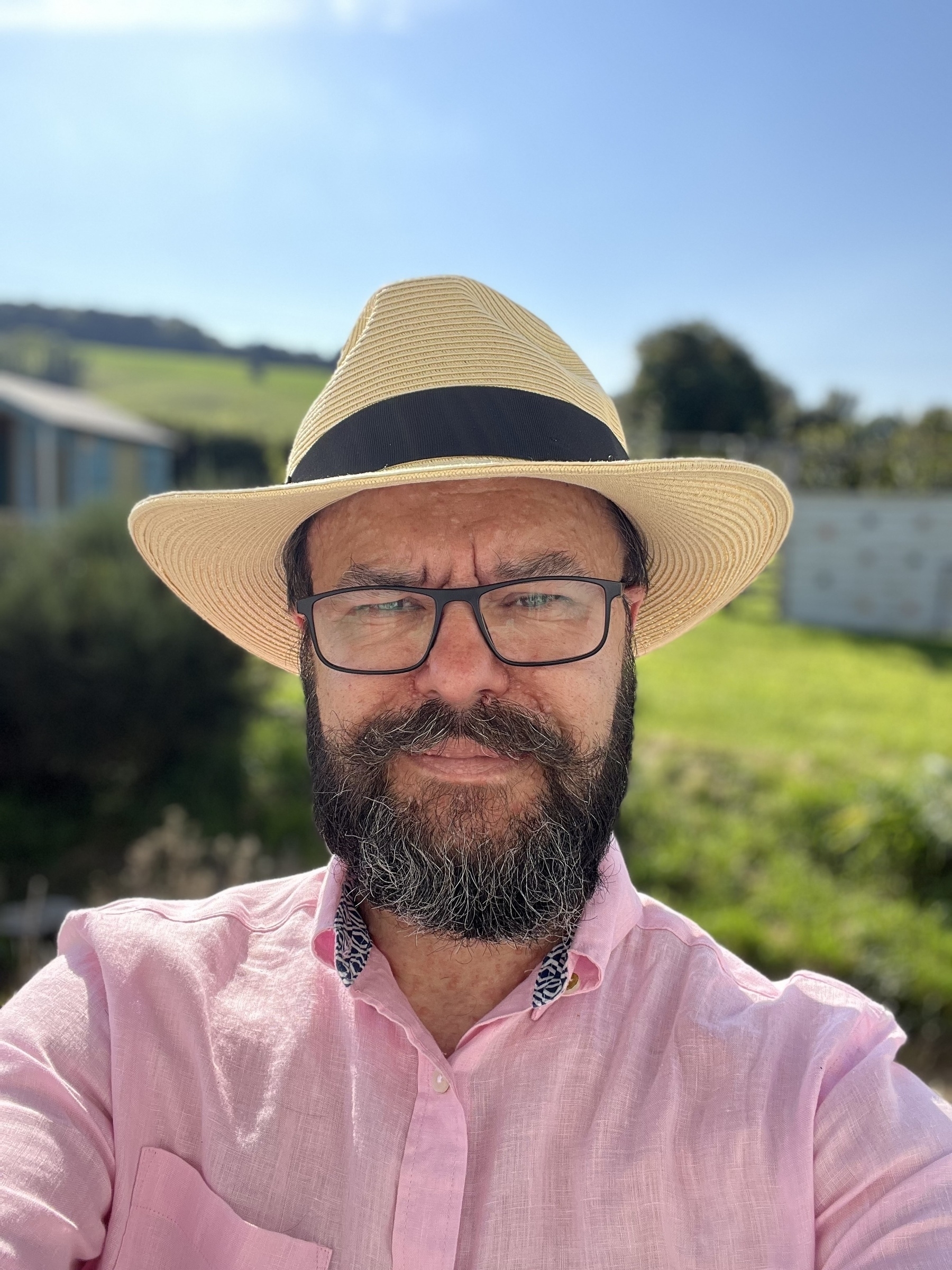 A beardy faced man grins smugly. He has glasses, wears a pink linen shirt and a straw fedora sits atop his huge bonce. 