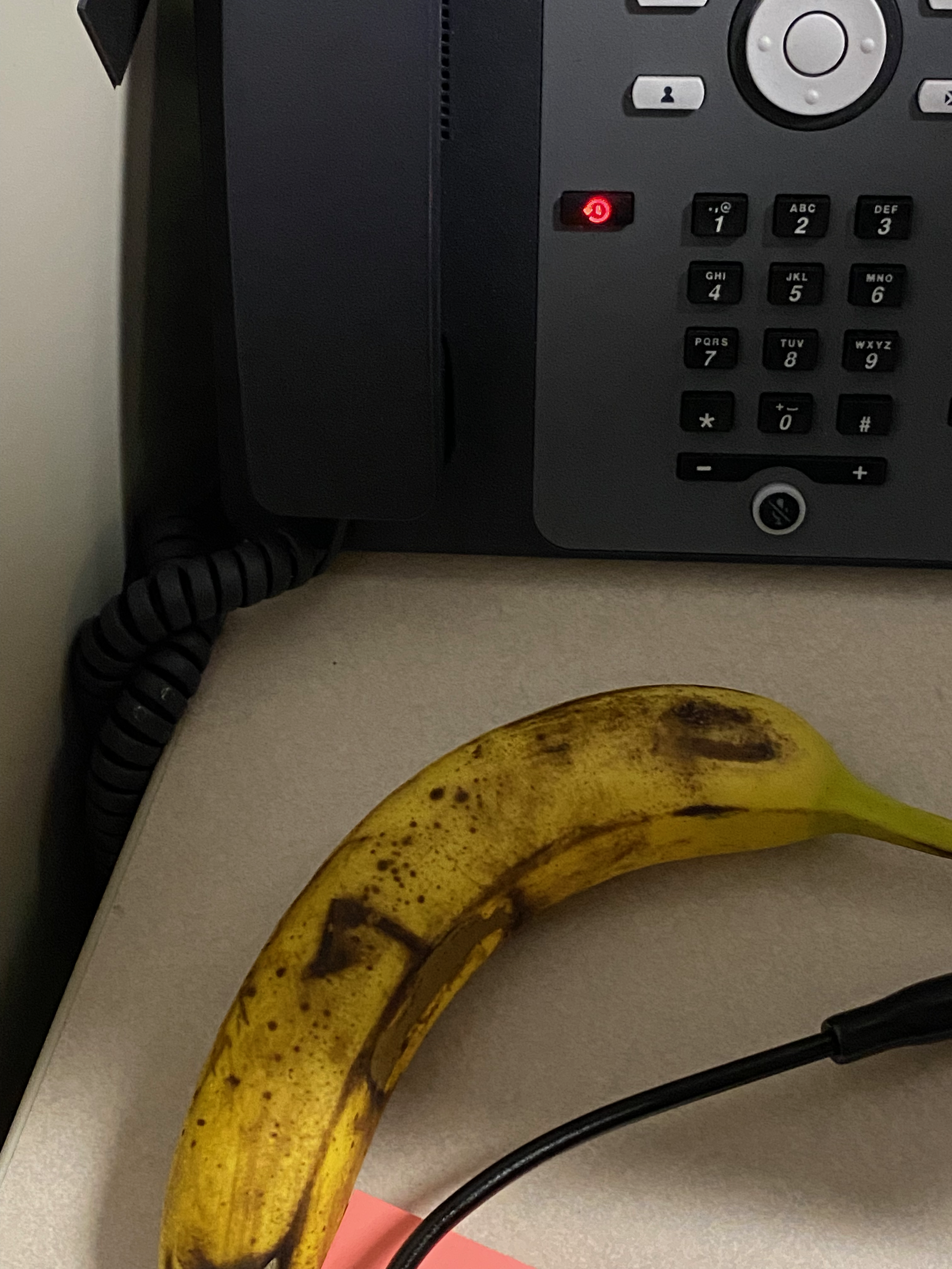 It's a picture of a ripe looking banana in front of a phone beside a non-descript black cord. The corner of a pad of sticky notes is visible at the bottom edge of the frame.