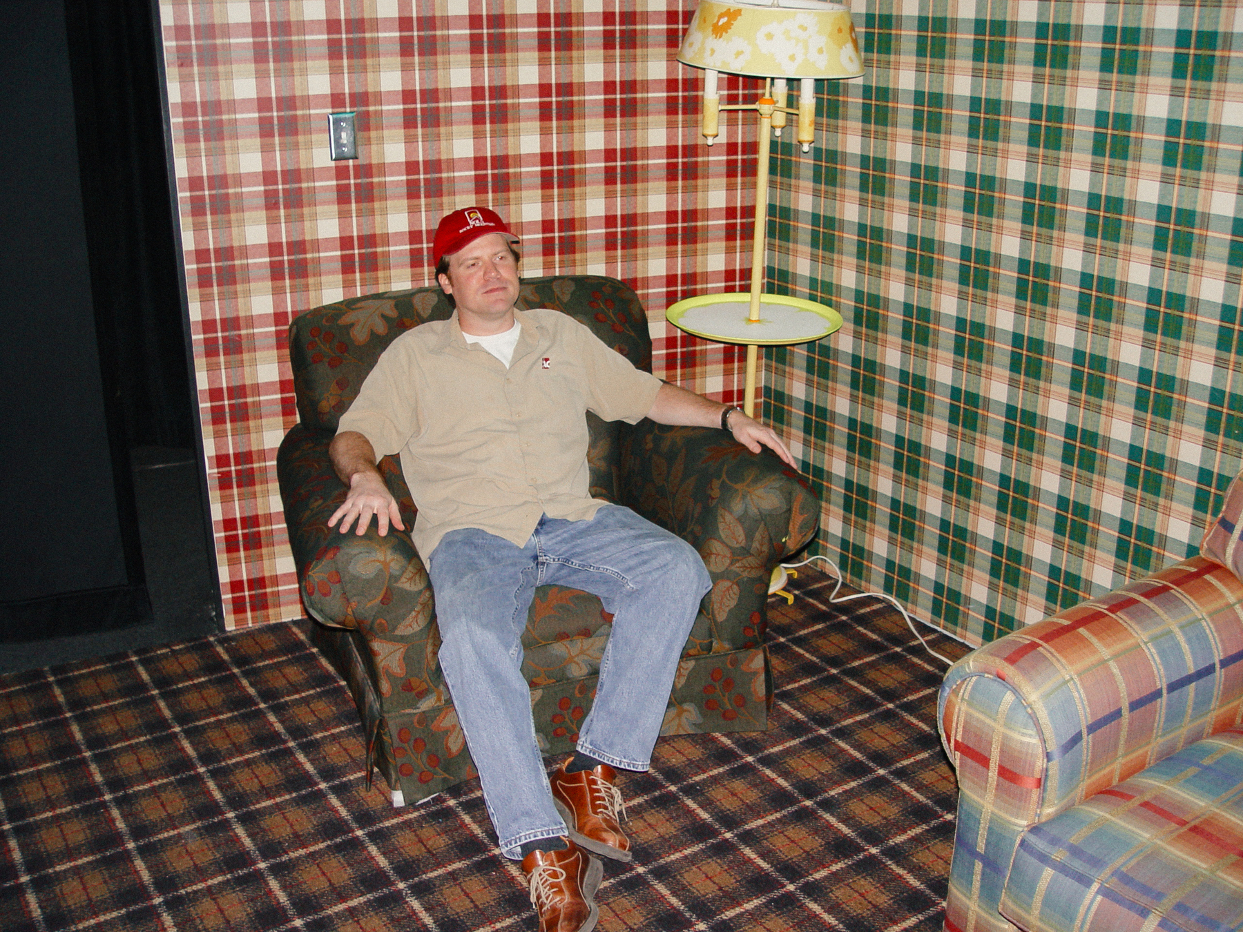 Man sits in stuffed arm chair in a set with different plaid wallpapers and tartan floor - Sony Cybershot