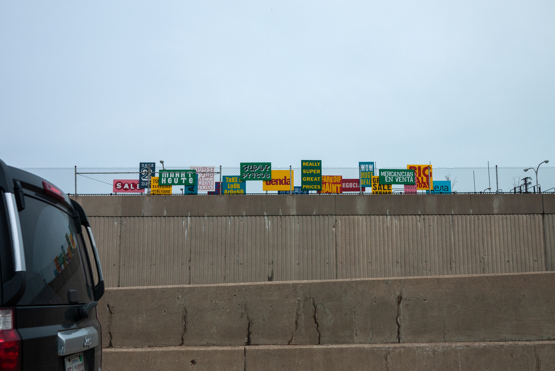 Colorful signs along a highway sound barrier that read "Sale, Super Prices, Tienda, Really Super Great Prices and the like - Panasonic Lumix GF1