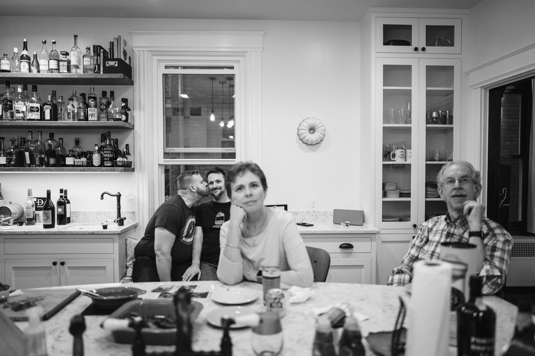 A man smootches his husband in the background of a kitchen while his parents look at the camera seated at the island. They're out of focus. - Leica Q