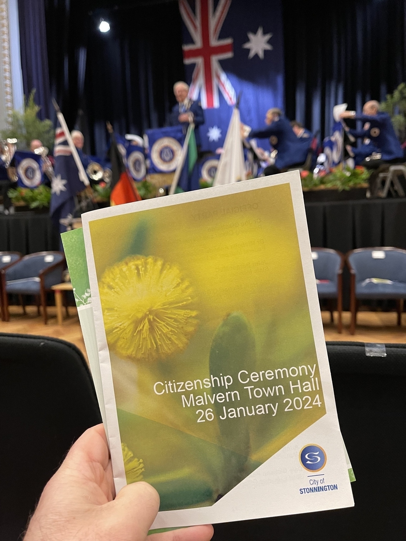In the foreground a pamphlet titled ‘Citizenship Ceremony’ and the background a traditional brass band.