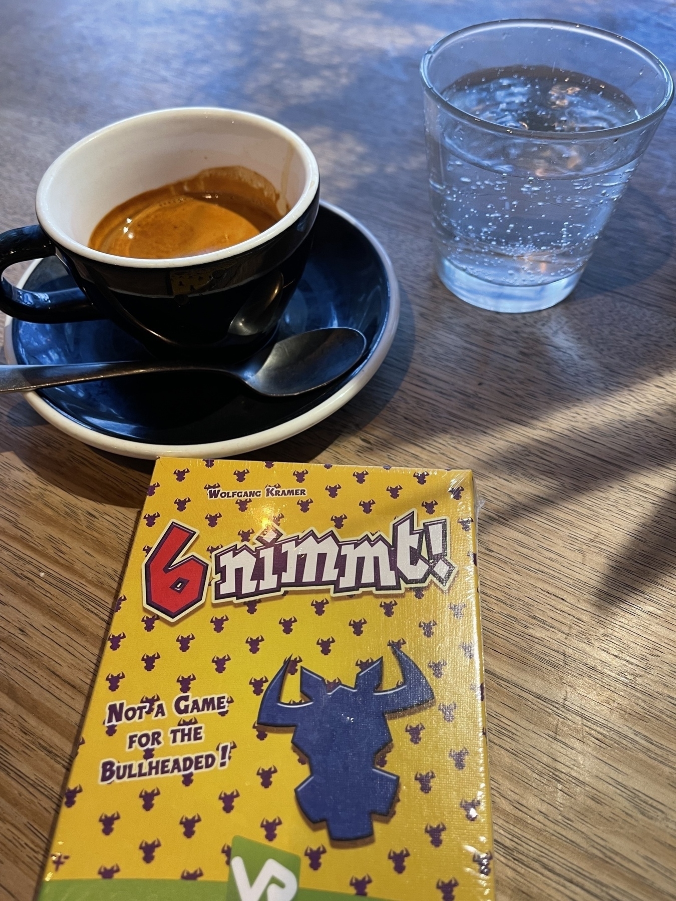 In the foreground a box of ‘6 nimmt’ (Take 6) card game and in the background espresso coffee in a blue cup and saucer and a small glass of sparkling water.