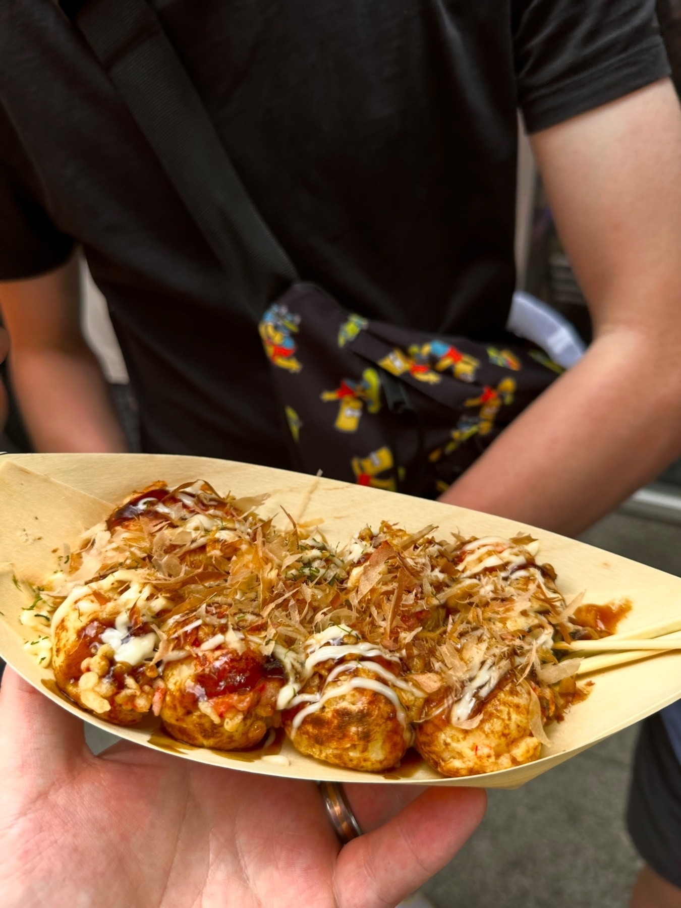 A plate of takoyaki, which are Japanese octopus dumplings, topped with mayonnaise, a savory sauce, and bonito flakes, held by a person with chopsticks.