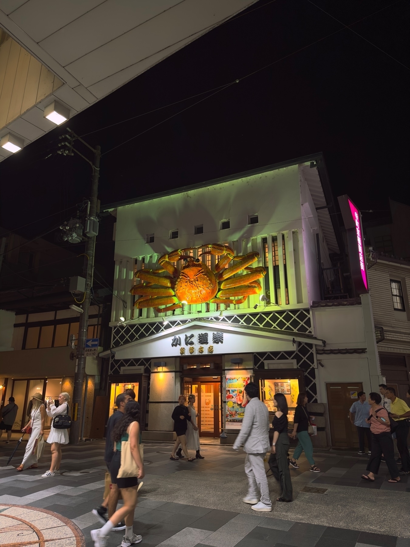 Nighttime street view of a building with a large illuminated 3D crab sculpture above the entrance, people walking by on the sidewalk, streetlights, and signage with Japanese characters.