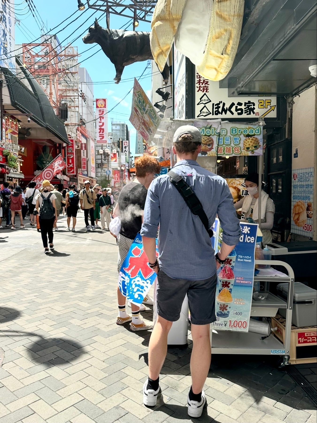 A busy street scene with pedestrians, a hanging sculpture of a cow, oversized model food items, and various signage in the shopping district of Osaka, Japan.