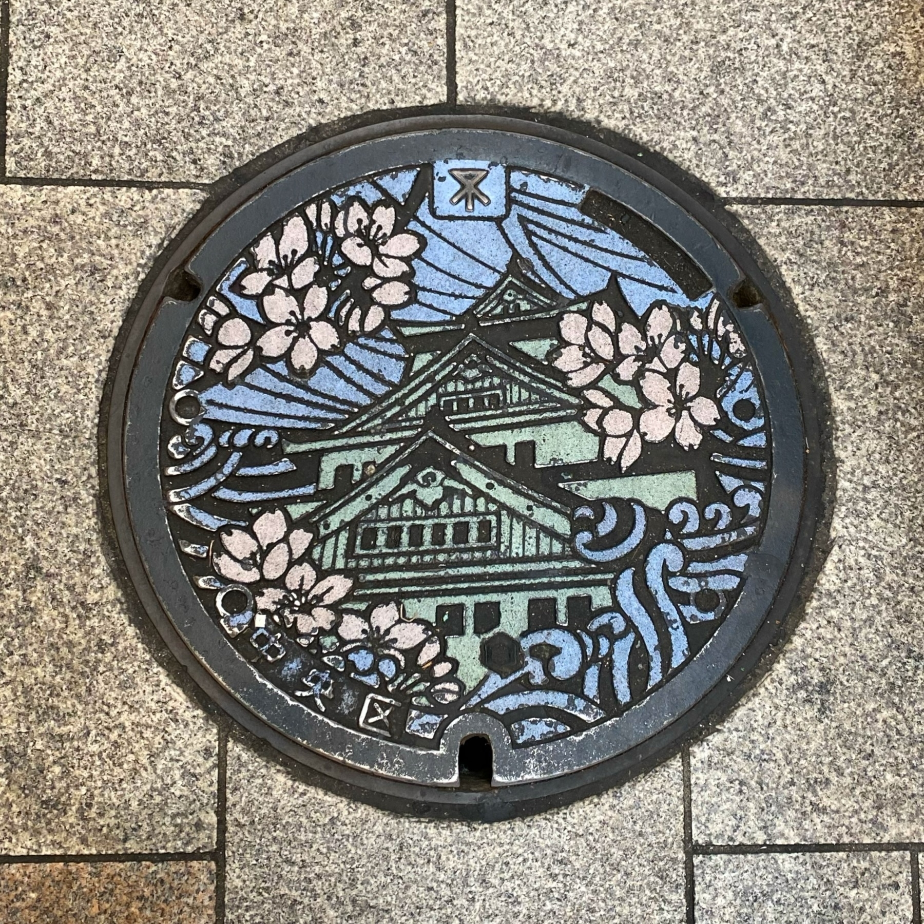 Decorative manhole cover featuring a traditional building, cherry blossoms, and stylized clouds or waves, showcasing Japanese cultural motifs.