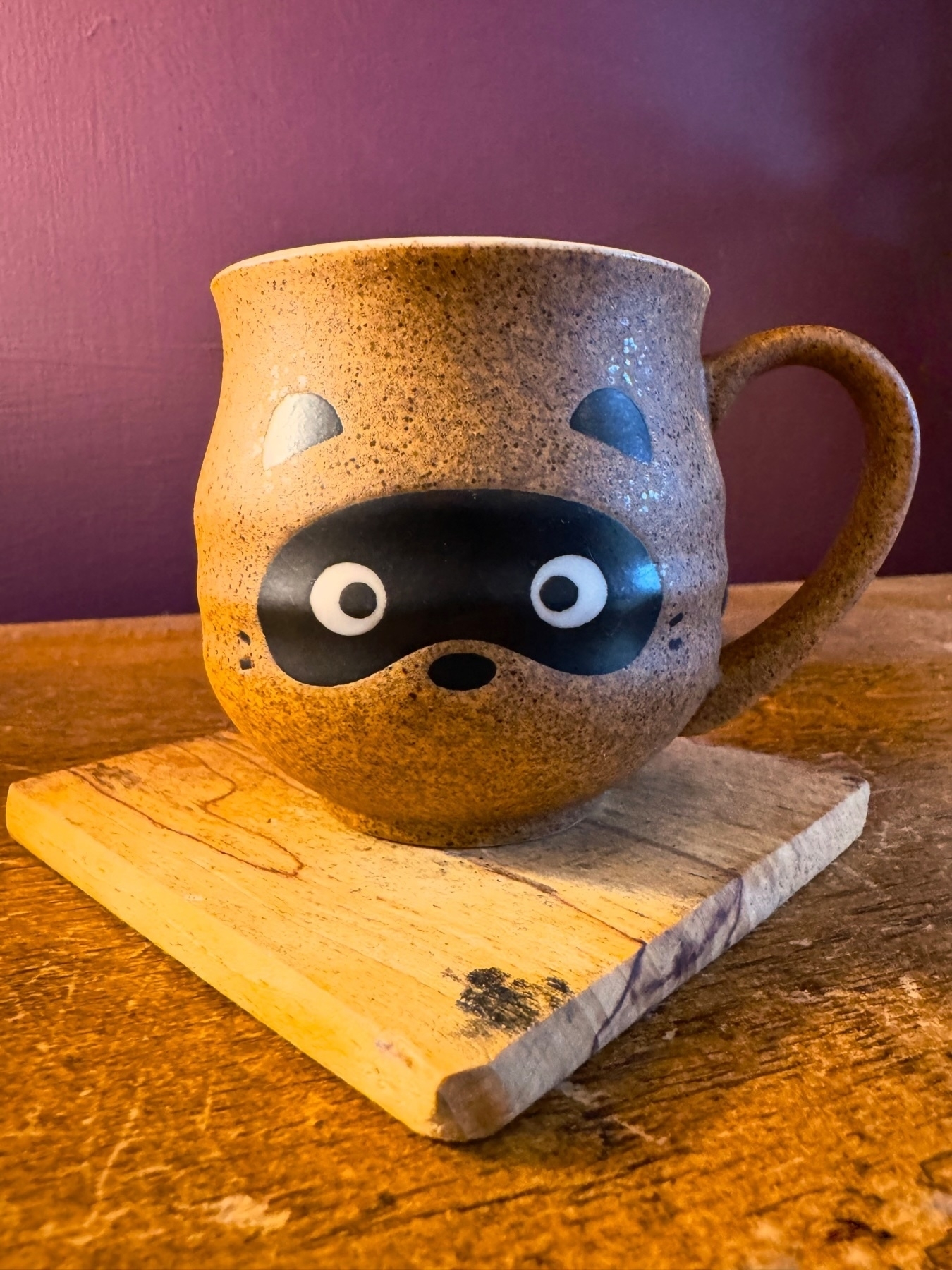 A ceramic mug designed to look like a raccoon face, placed on a wooden coaster on a wooden table with a purple wall background.