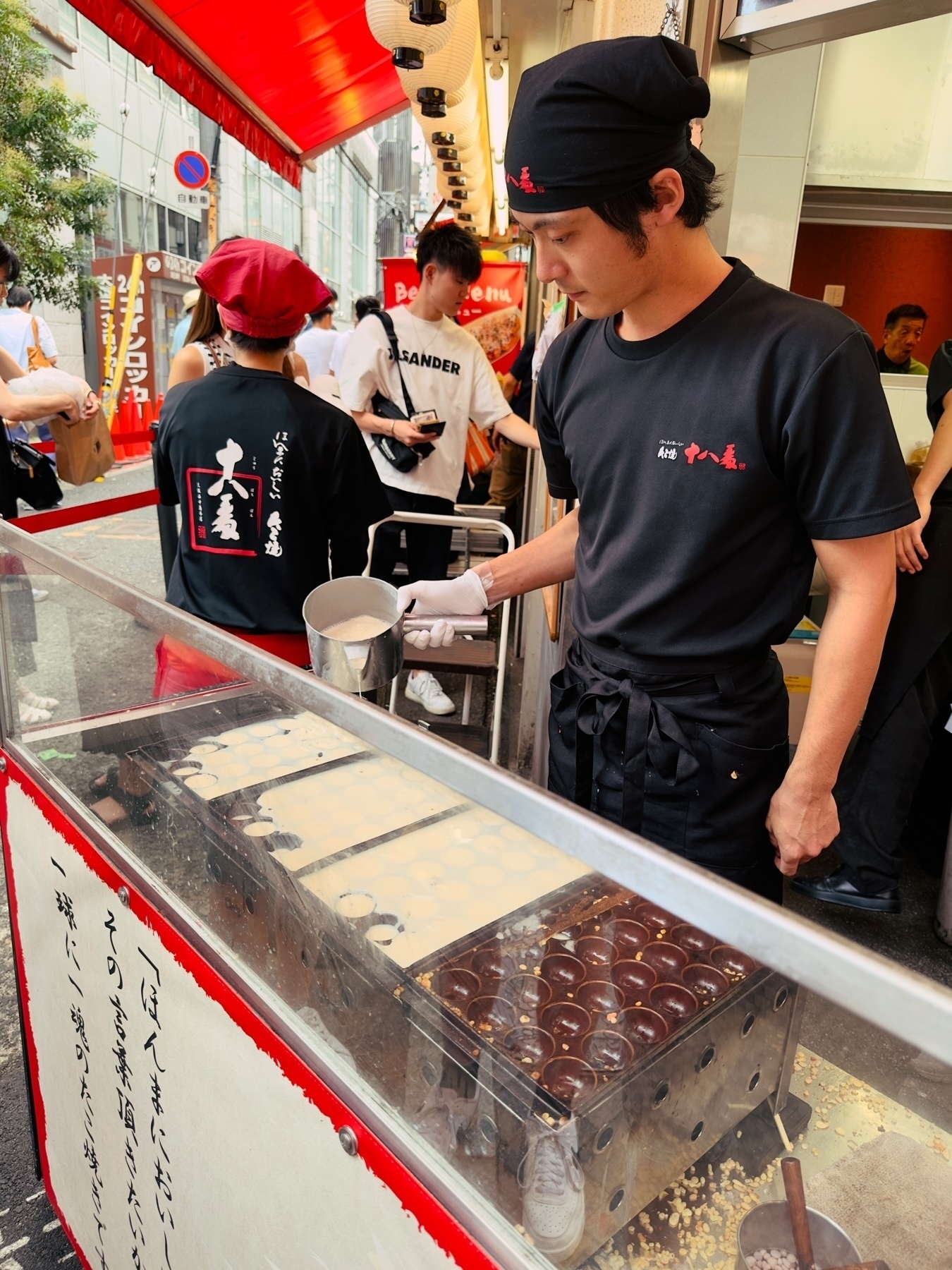 A street vendor in a black T-shirt and chef hat preparing food at a stall with a glass display showcasing rows of round, light-colored dough balls. Behind him, pedestrians and shops can be seen under a red awning.