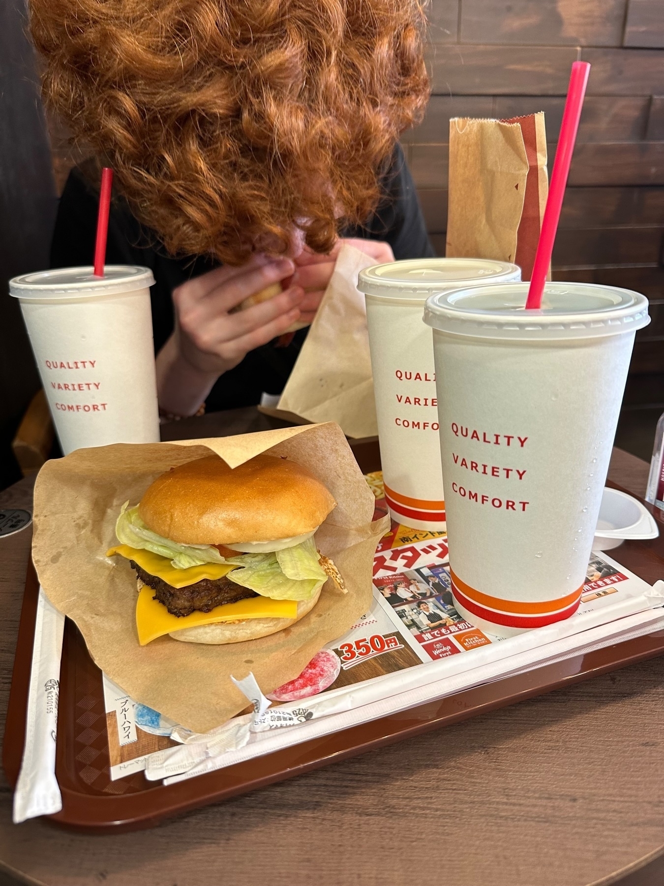A person with curly red hair eating a sandwich, a cheeseburger with lettuce on a tray, two drinks with red straws, disposable cutlery, and fast food restaurant branding.