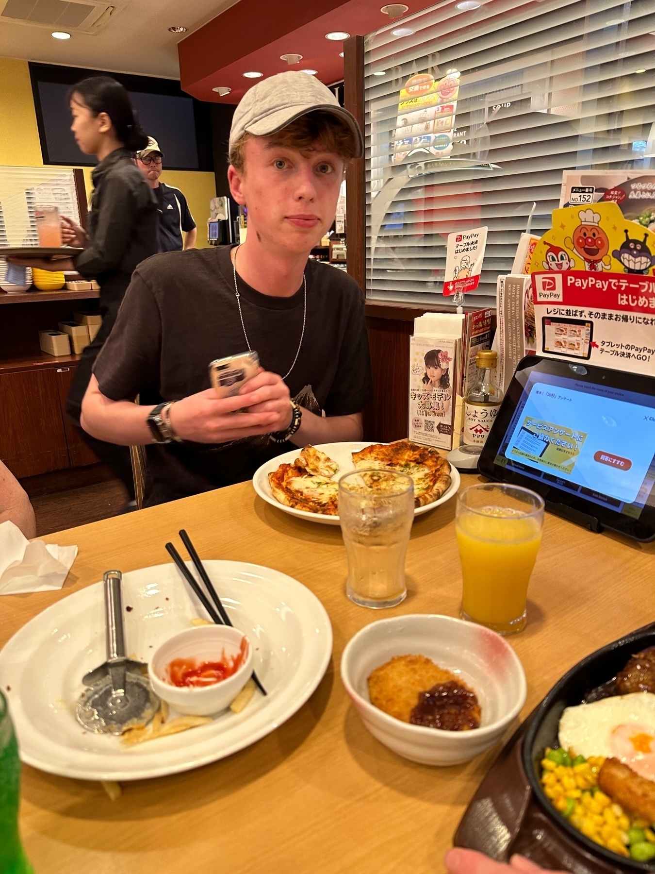 My son sitting at a restaurant table with various dishes including pizza, eggs, and corn, holding a can with a surprised expression.