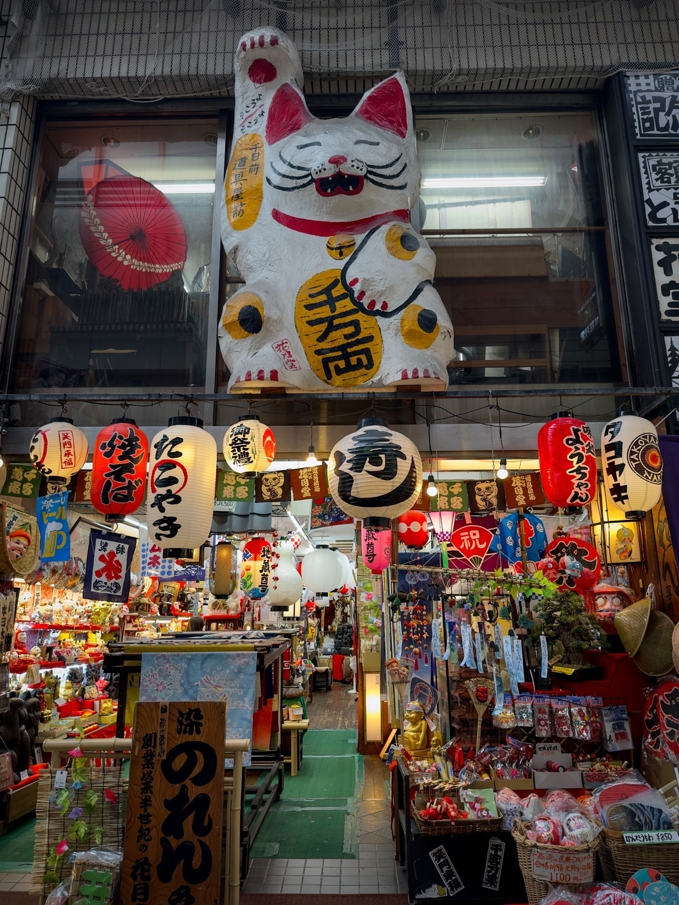 This image shows a Japanese market alley with traditional goods and decorations, featuring a large Maneki-neko (lucky cat) statue, colorful lanterns, banners, and various cultural and souvenir items on display.