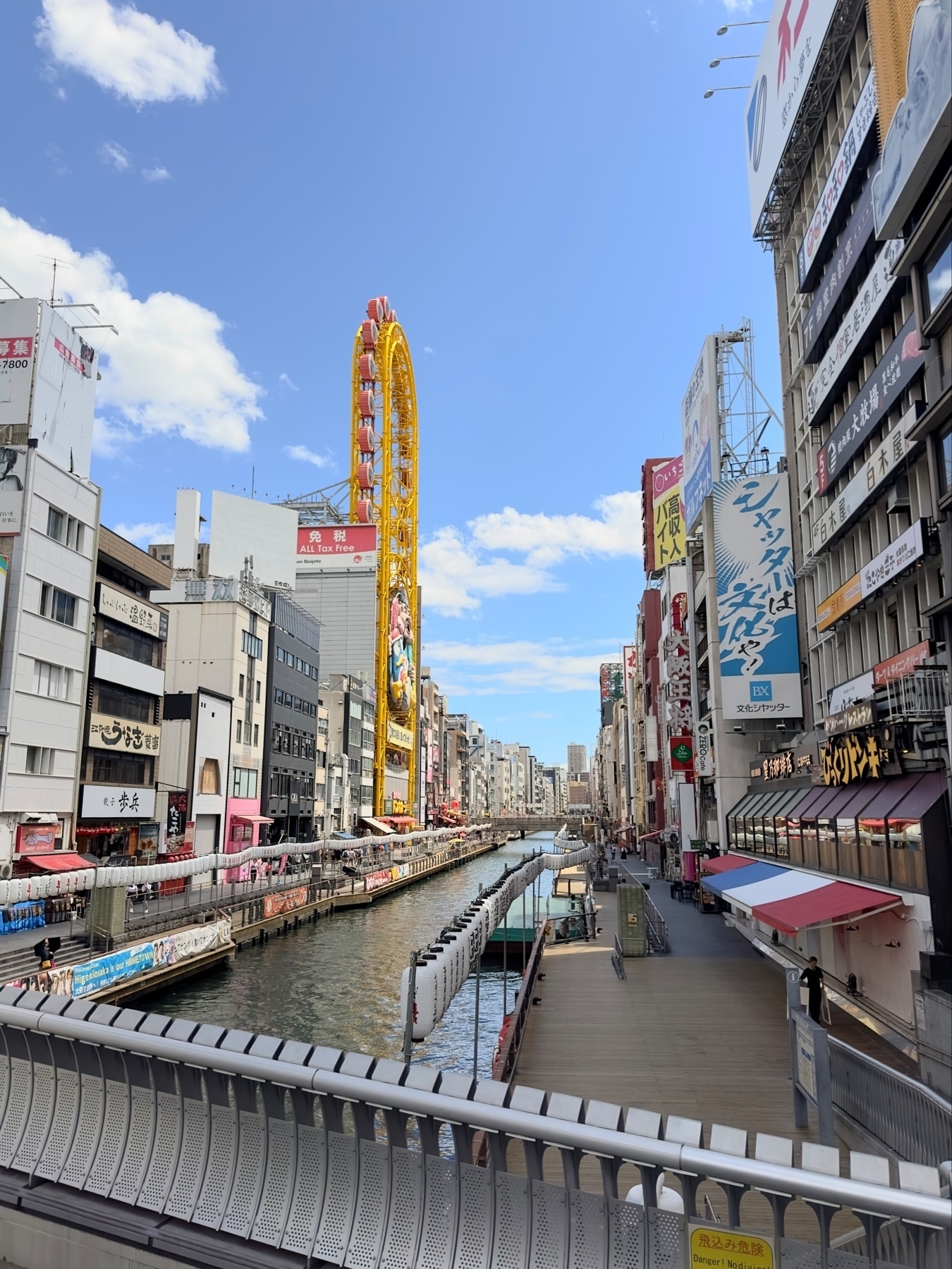 A vibrant urban street scene in Japan with a large Ferris wheel, colorful billboards, and a canal running through it, flanked by buildings and pedestrian walkways.