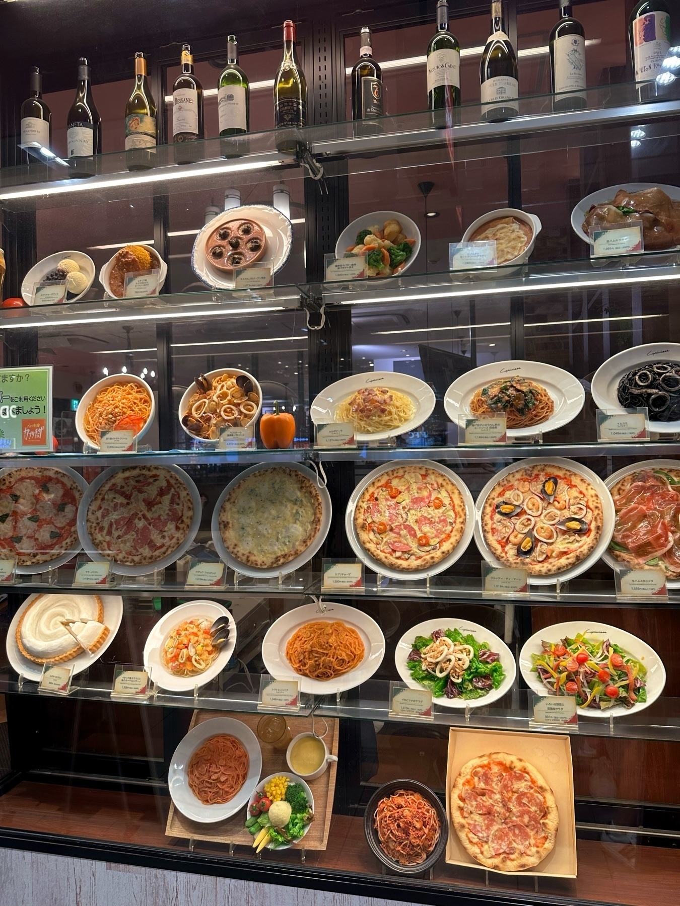 Display of various Italian dishes including pasta, pizza, and salad, alongside bottles of wine at the top shelf, likely in a restaurant or food establishment. Some dishes have descriptive tags in front.