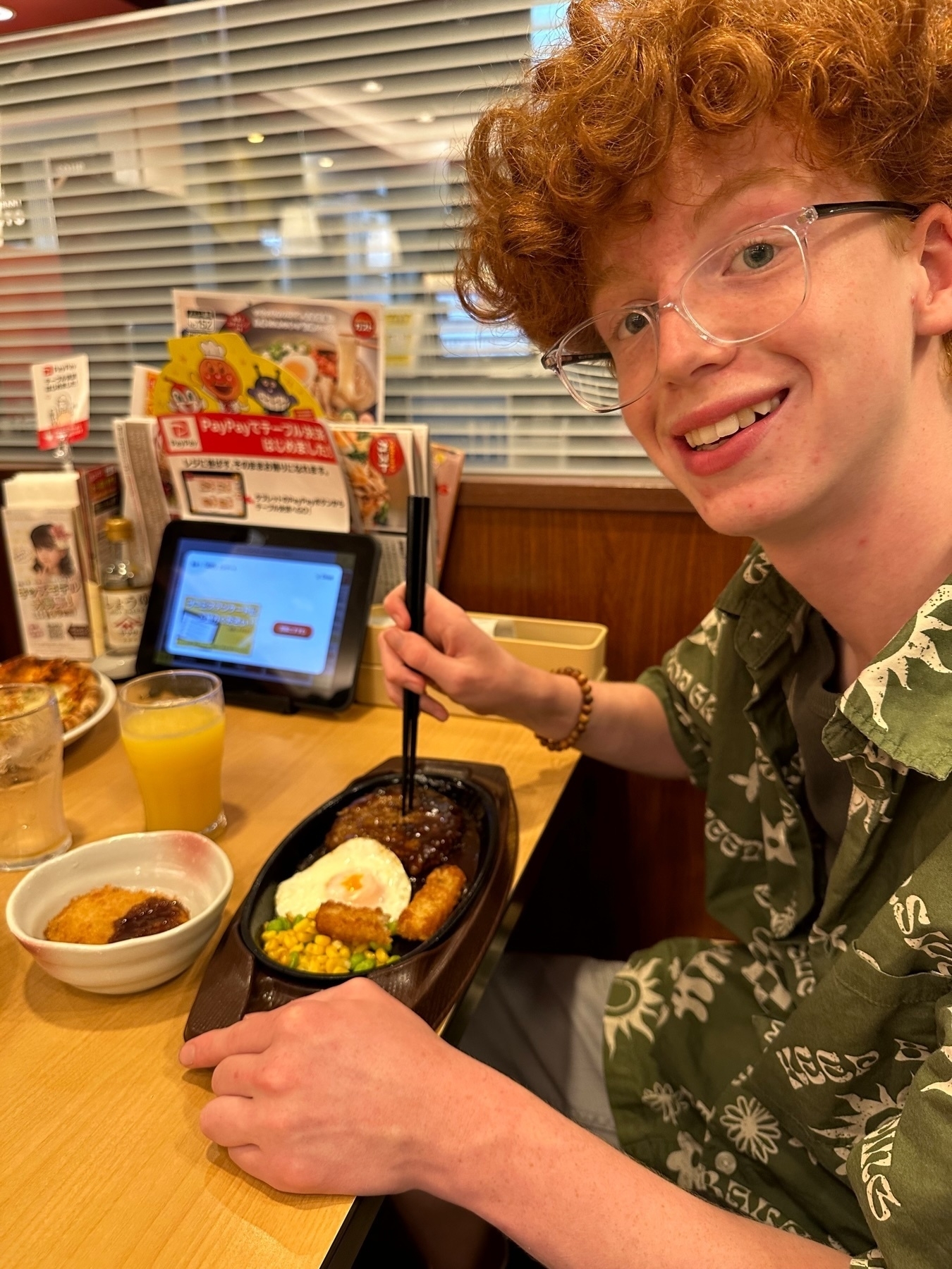 A smiling person with red curly hair, wearing glasses and a green patterned shirt, seated at a restaurant table with a tray of food including a hamburger steak with an egg on top, hash browns, and corn, with a side of ketchup