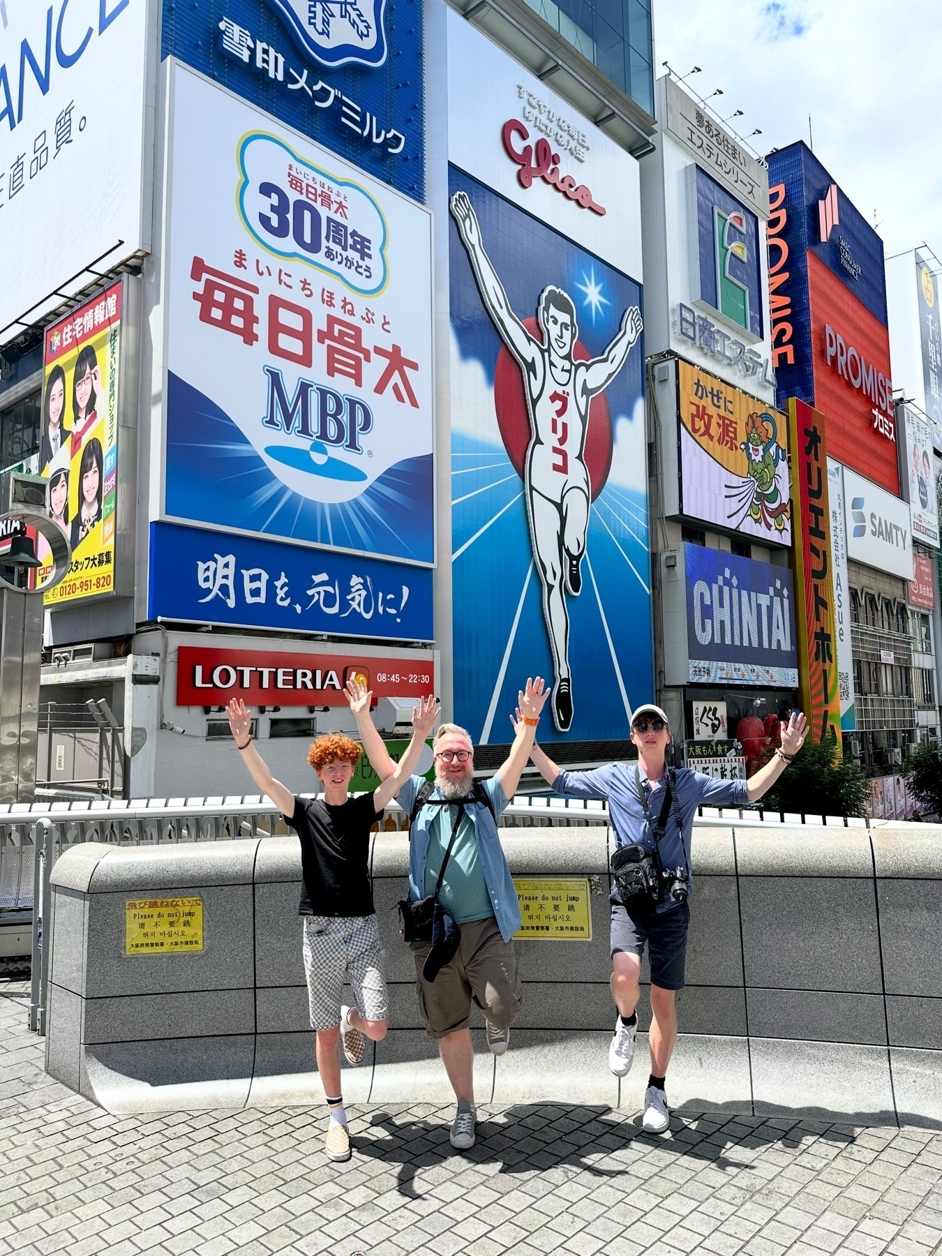 Me and my sons joyfully posing with raised arms in front of the famous Glico Running Man sign in the Dotonbori district of Osaka, Japan, with various colorful billboards in the background.