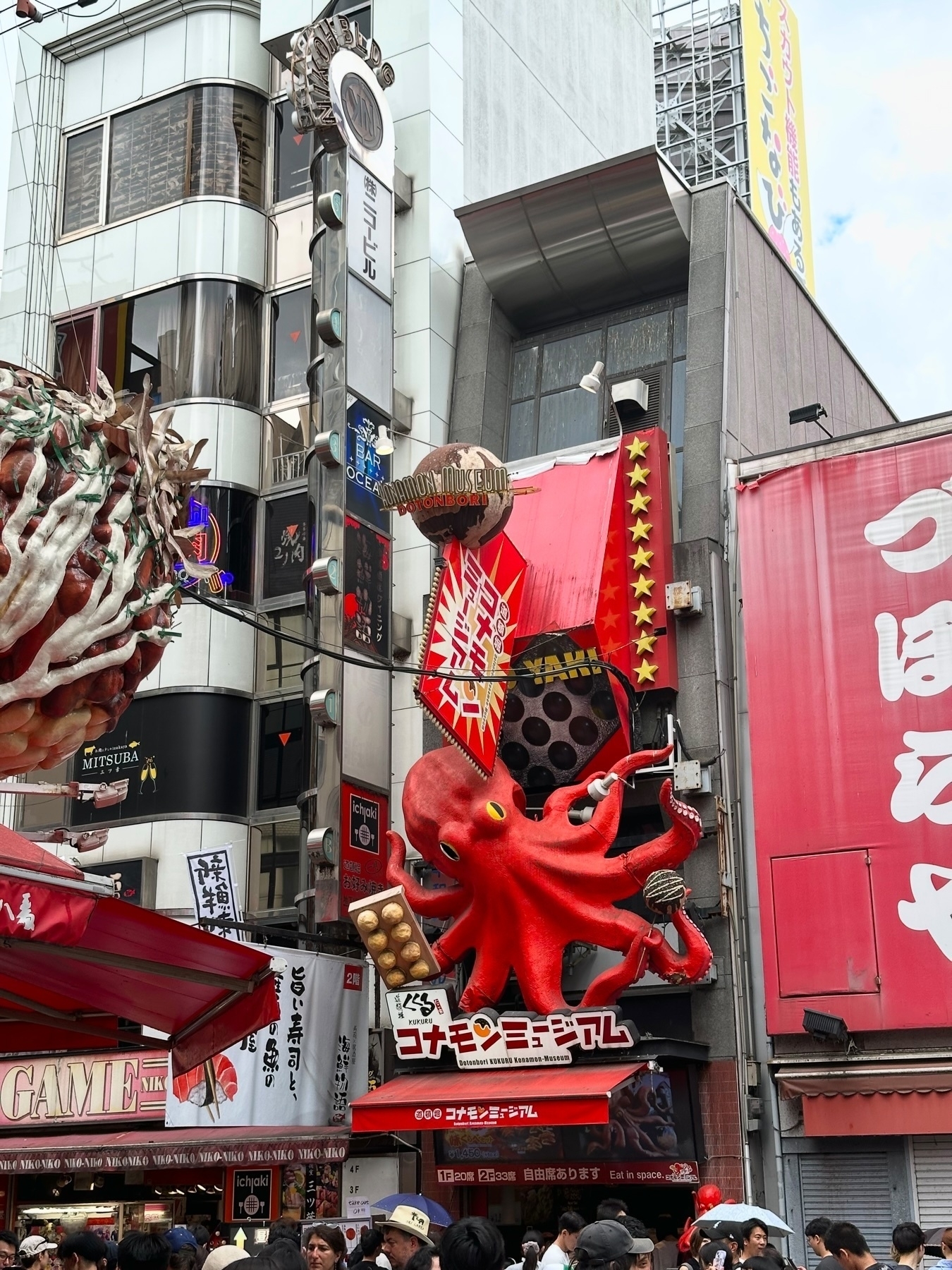 A busy street scene in Japan with a large red octopus sculpture above a restaurant entrance, surrounded by various signs and banners, and a crowd of people walking below.