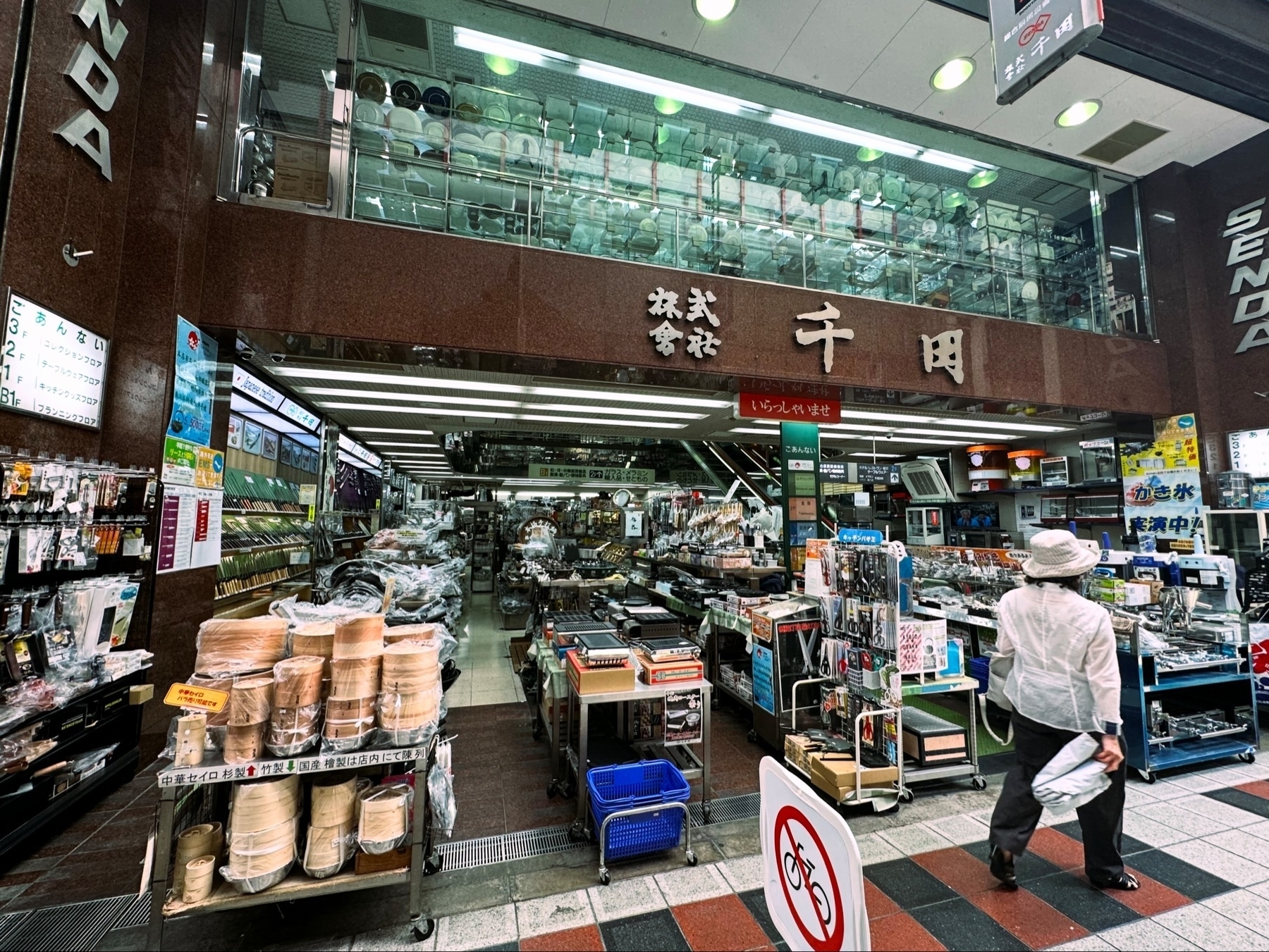 A person walking past the entrance of an electronics store, which displays a variety of products ranging from kitchenware to electronic equipment. Visible signage includes Japanese text.