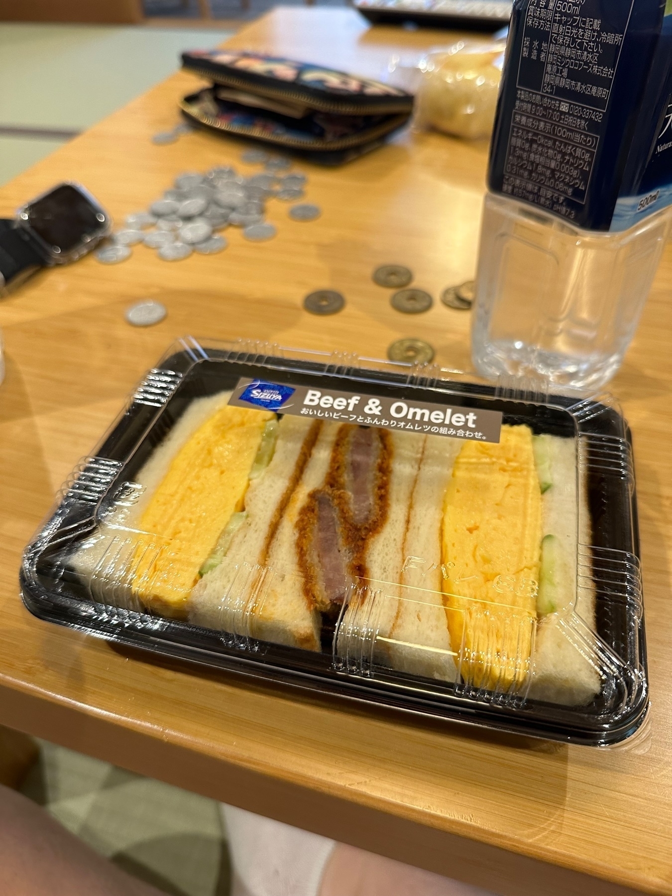 A Beef &amp; Omelet sandwich in a clear plastic container on a wooden table with scattered coins, a wallet, a smartwatch, and a partially visible water bottle in the background.