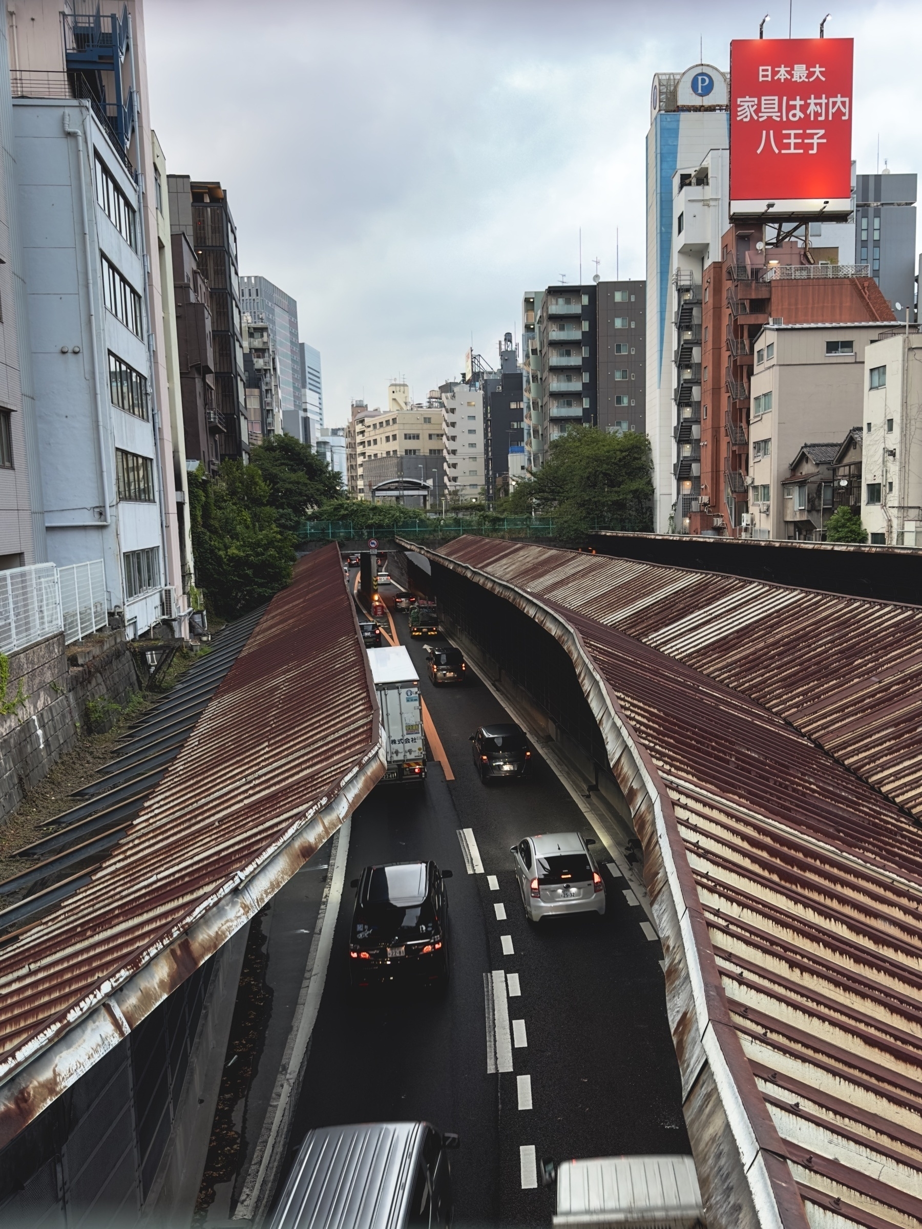 A Japanese city road surrounded by tall buildings with a heavily rusted top over the road