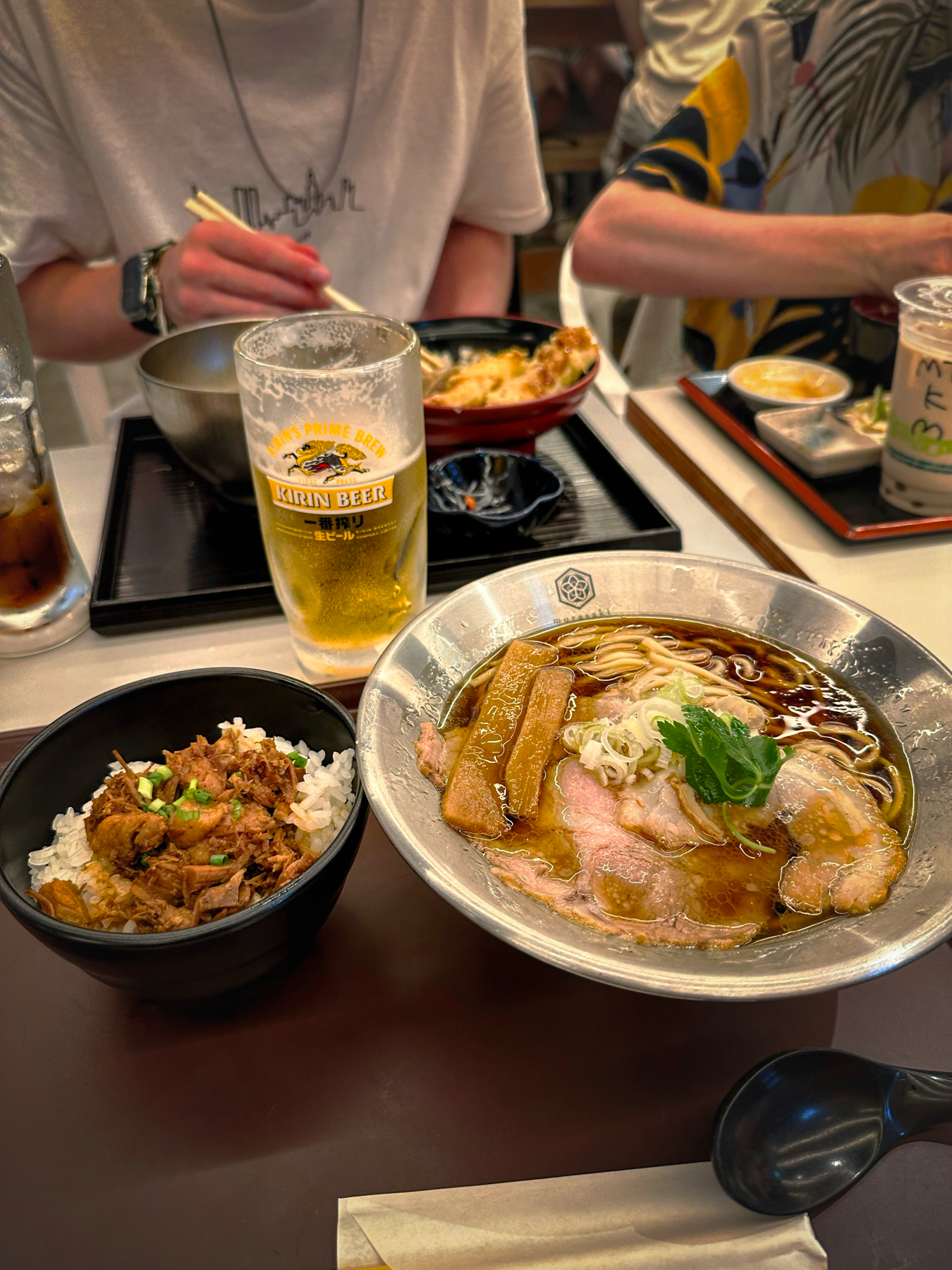 A table in a restaurant with Japanese dishes, including a bowl of ramen with sliced pork and a rice bowl with beef topping. There is also a glass of Kirin beer and two people dining in the background.