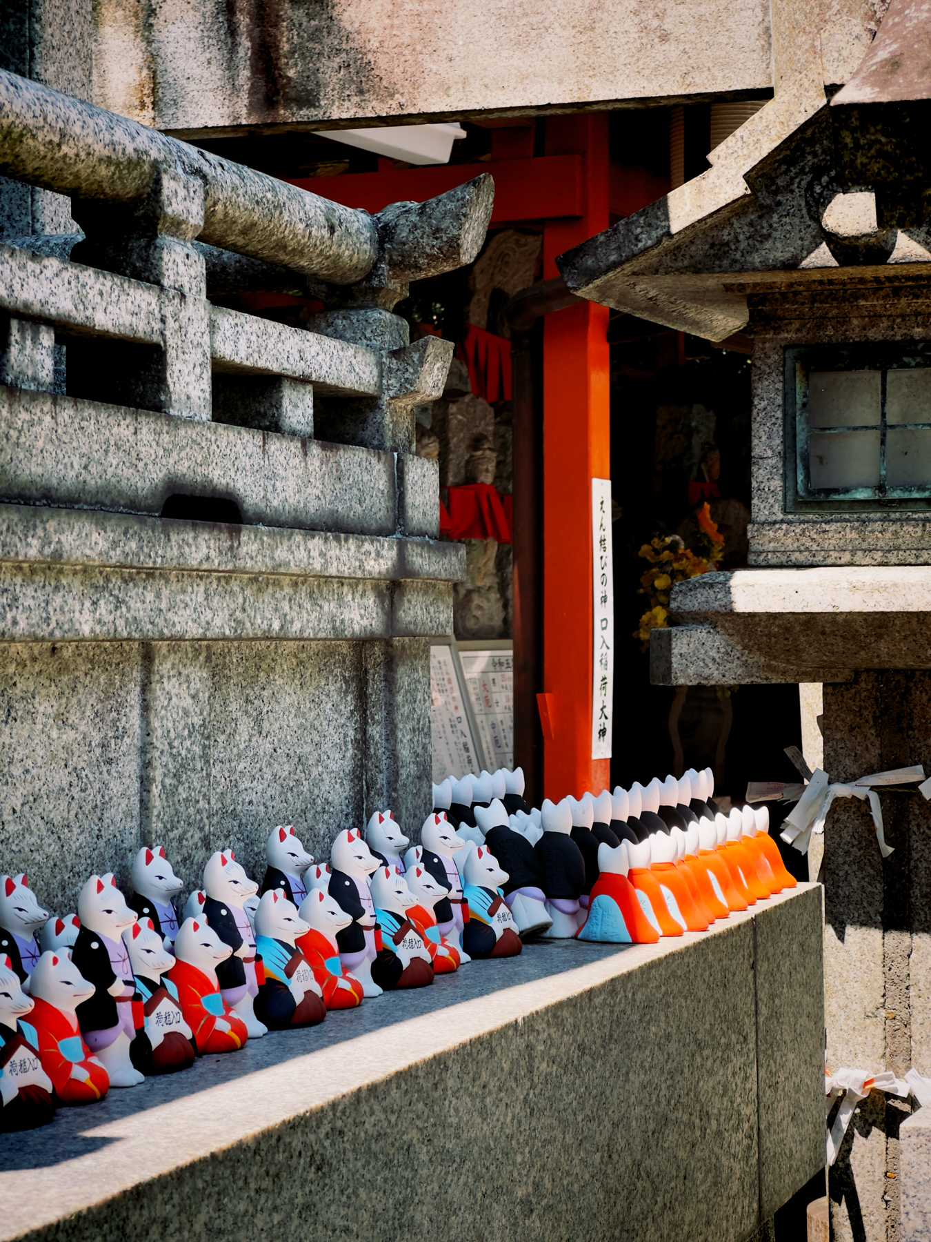 A collection of traditional Japanese fox statues known as &ldquo;Inari,&rdquo; in various colors, lined up at a Shinto shrine, with a torii gate and stone lantern visible in the background.