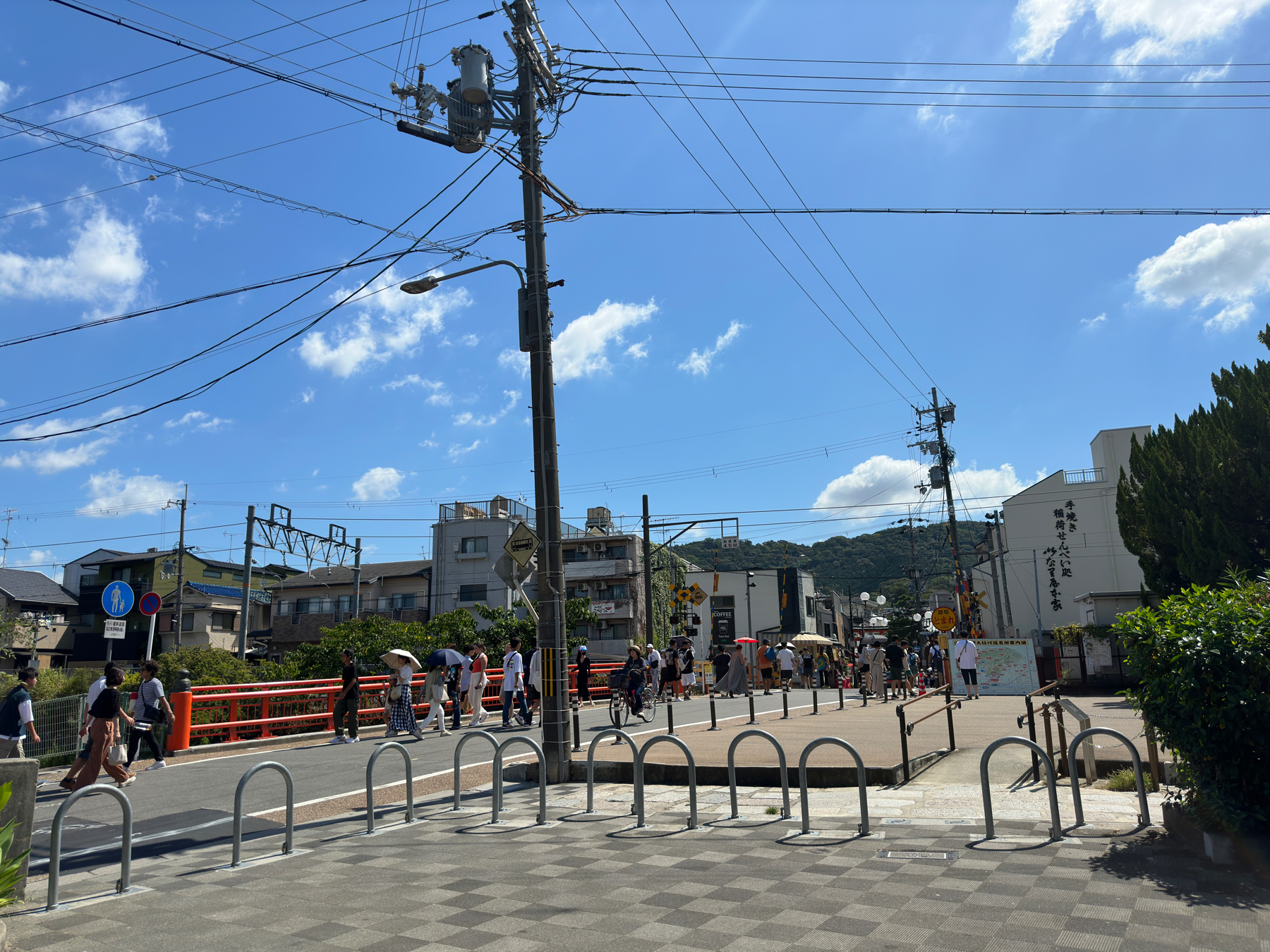 A sunny street scene with people walking near a red bridge, overhead power lines, bicycles racks, and various signage, including Japanese characters. There&rsquo;s a mix of modern buildings and lush greenery in the background.