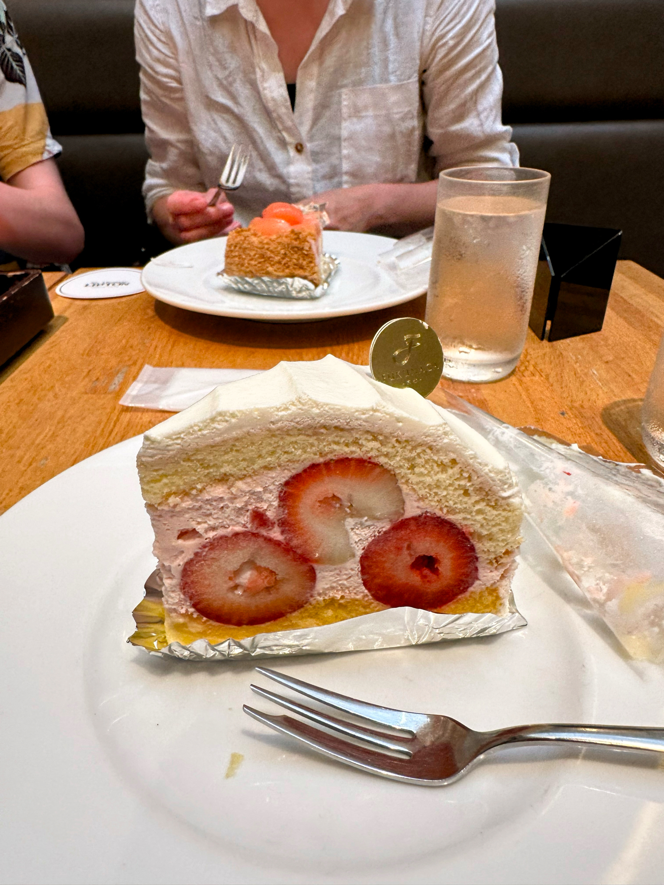 A piece of strawberry shortcake on a plate with a fork, featuring sliced strawberries in its filling, accompanied by another slice of cake with melon balls on another plate in the background. There&rsquo;s a glass of water on the table.
