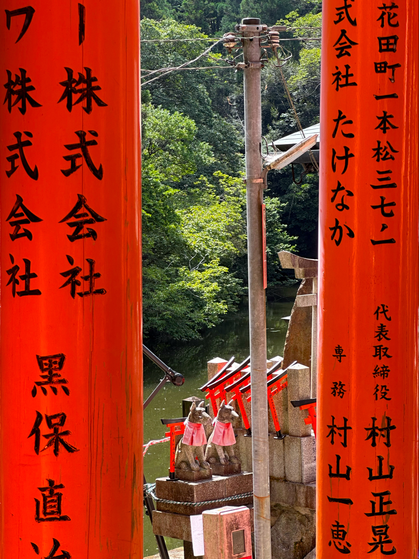 This image shows a pair of vibrant orange torii gates with black Japanese characters on them. Between the gates, a utility pole with wires is seen against a backdrop of lush green trees. Below, two traditional stone fox statues, dressed in red bibs