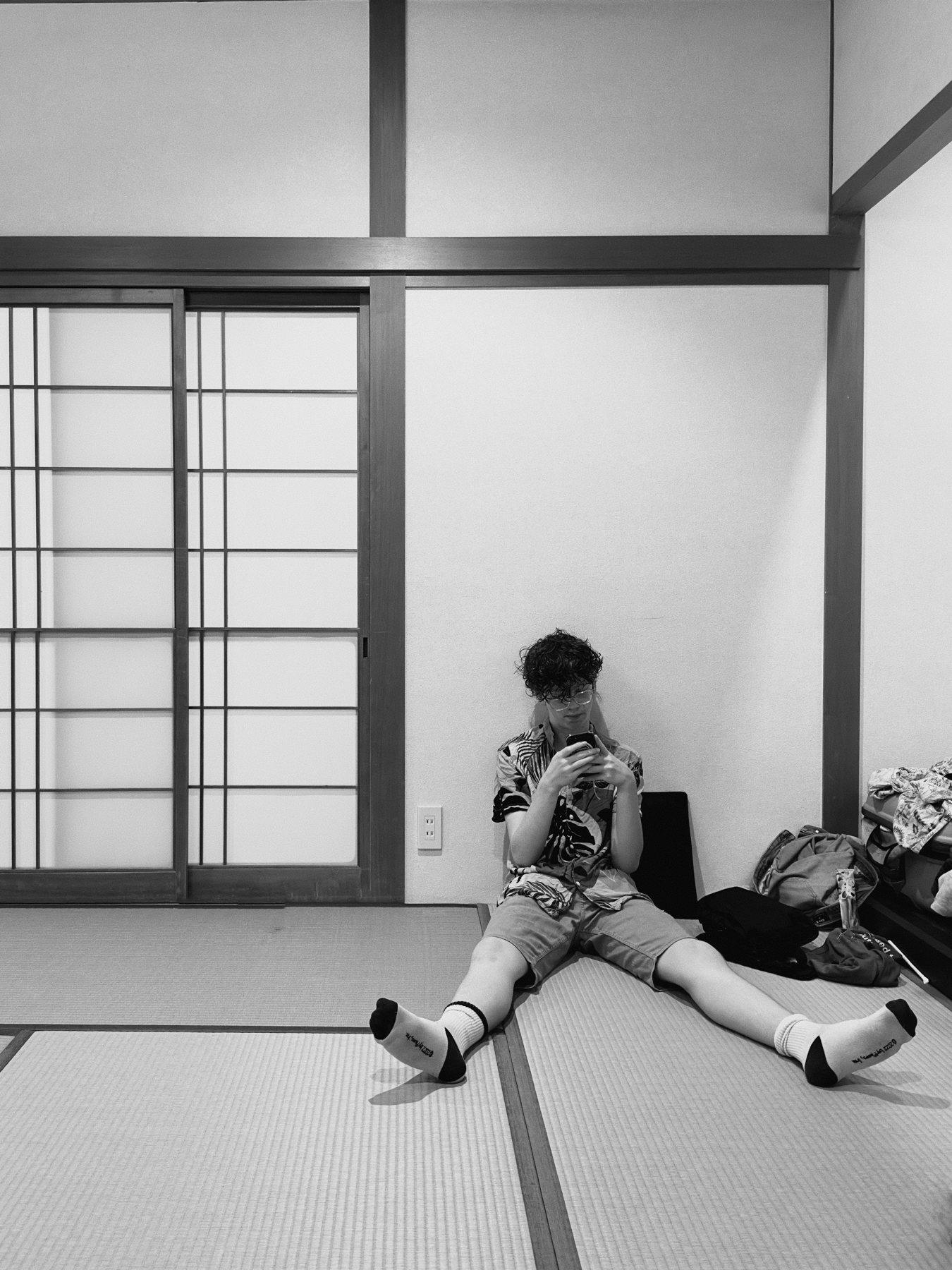 A person sits on tatami mats in a Japanese-style room, using a smartphone. There&rsquo;s a shoji sliding door to the side and belongings scattered next to the individual. The image is in black and white.