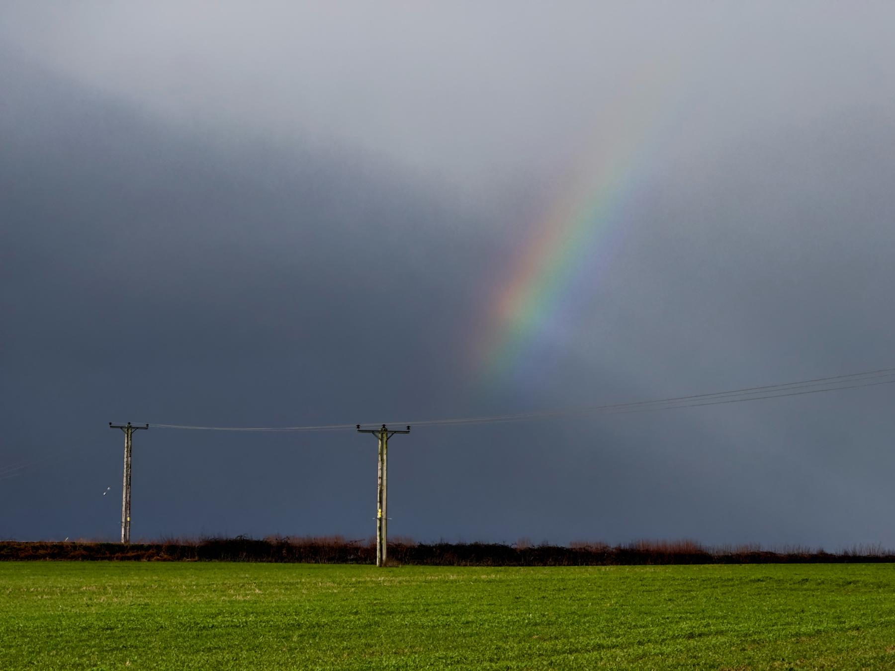 A landscape featuring a green field with two electricity poles and wires, with a partially visible rainbow in a dark cloudy sky.