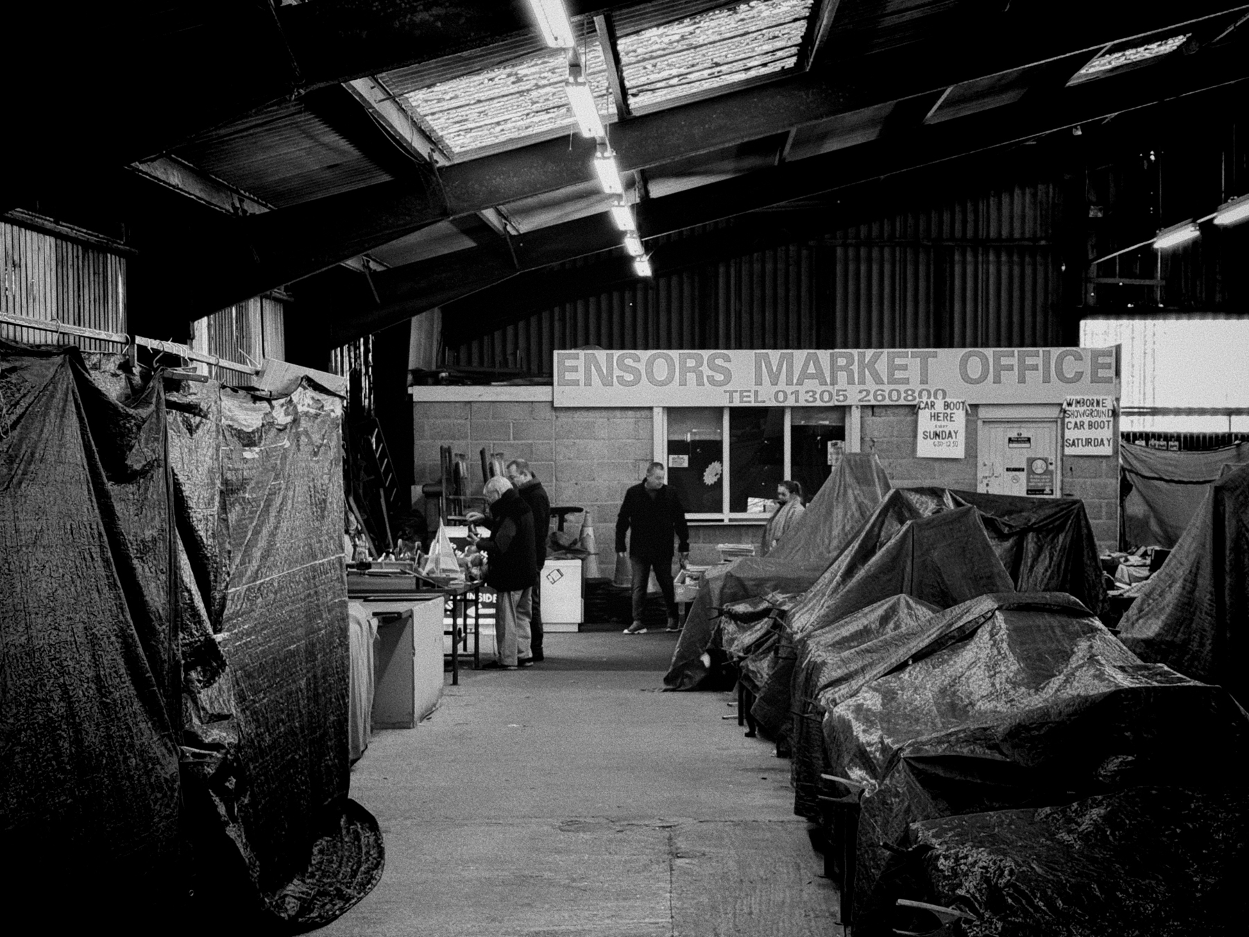 Black and white photo of an indoor market setting with covered stalls, a few individuals visible, and a sign reading &ldquo;ENSORS MARKET OFFICE&rdquo; with a telephone number.