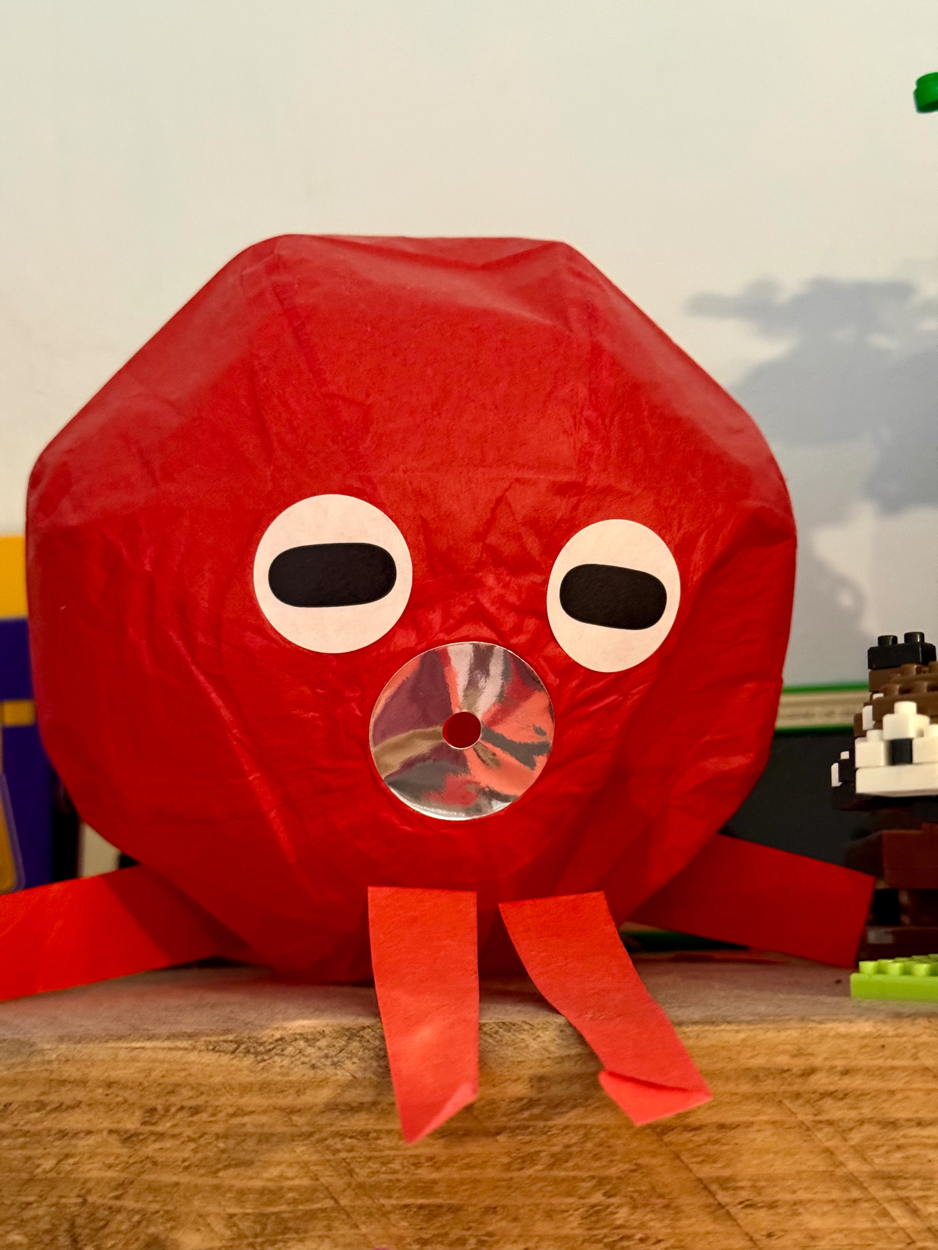 A red octopus crafted from paper materials, with white eyes, a circular clear plastic mouth, and tentacles arranged on a wooden surface.