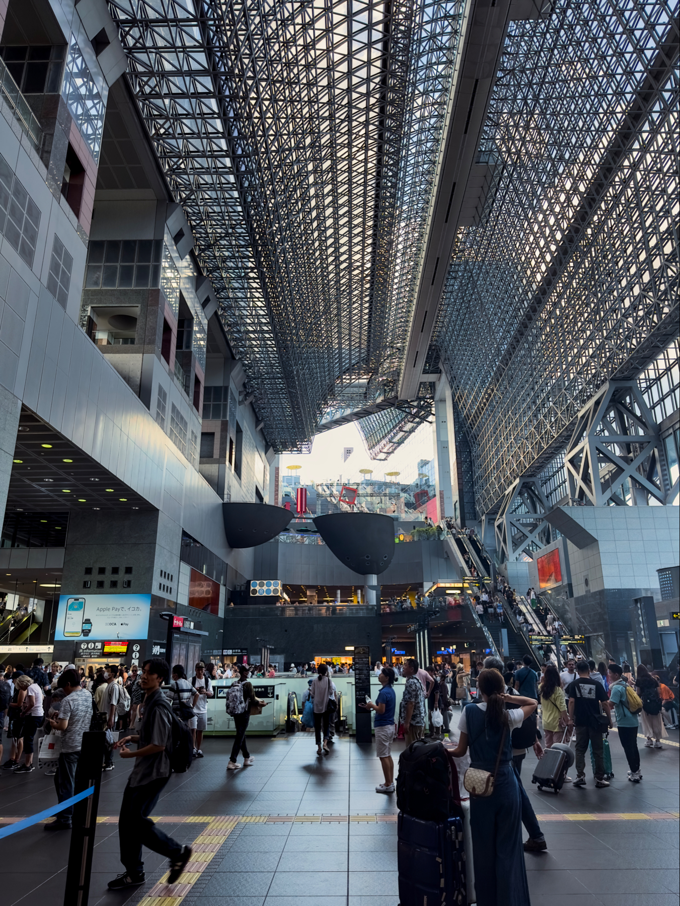 Interior of modern looking Kyoto train station with a high geometric ceiling, bustling with people and featuring escalators and large video screens.