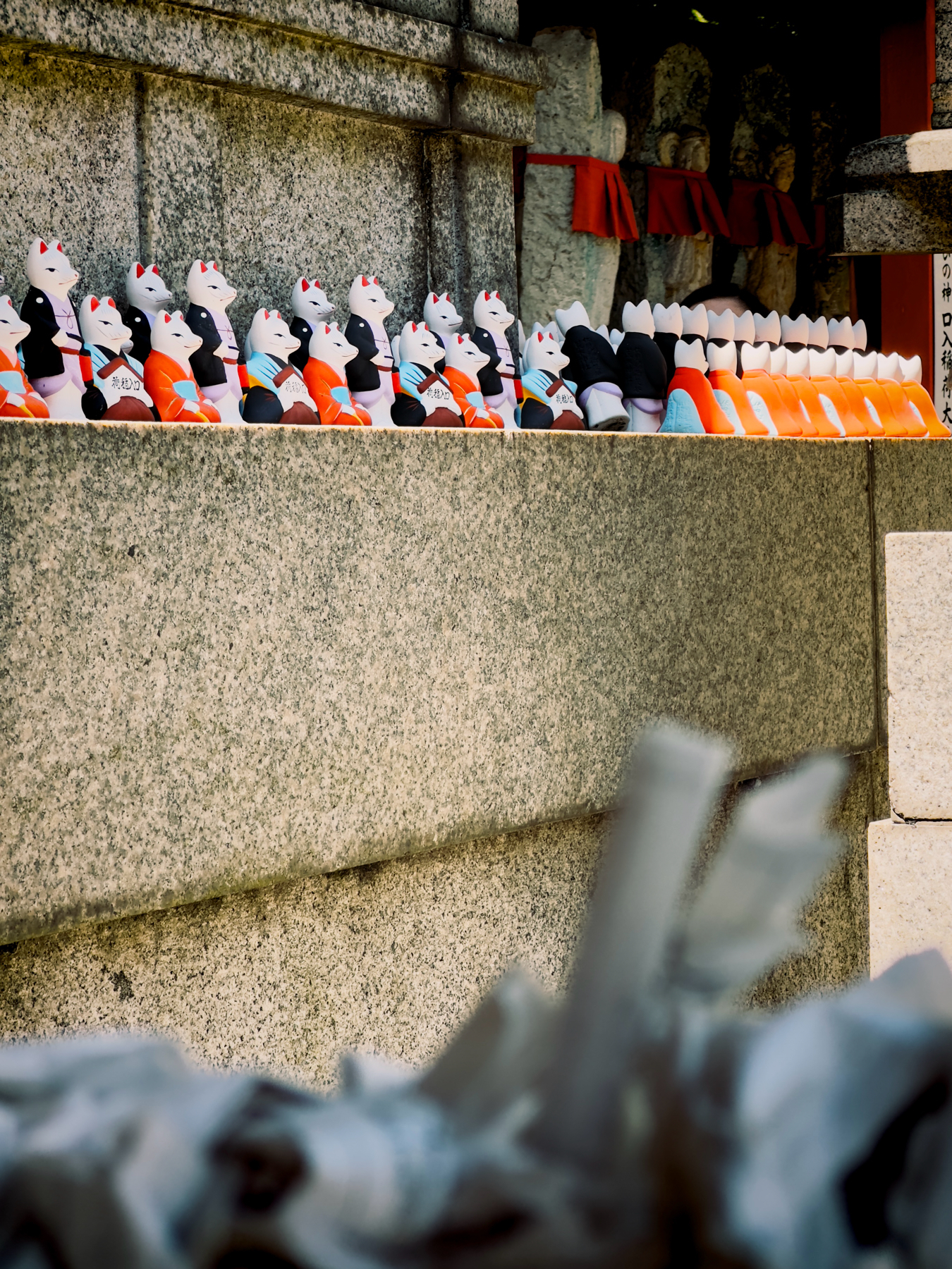 A series of traditional Japanese fox (kitsune) statues wearing red bibs, lined up on a shrine ledge with blurred foreground elements that resemble leaves or paper.