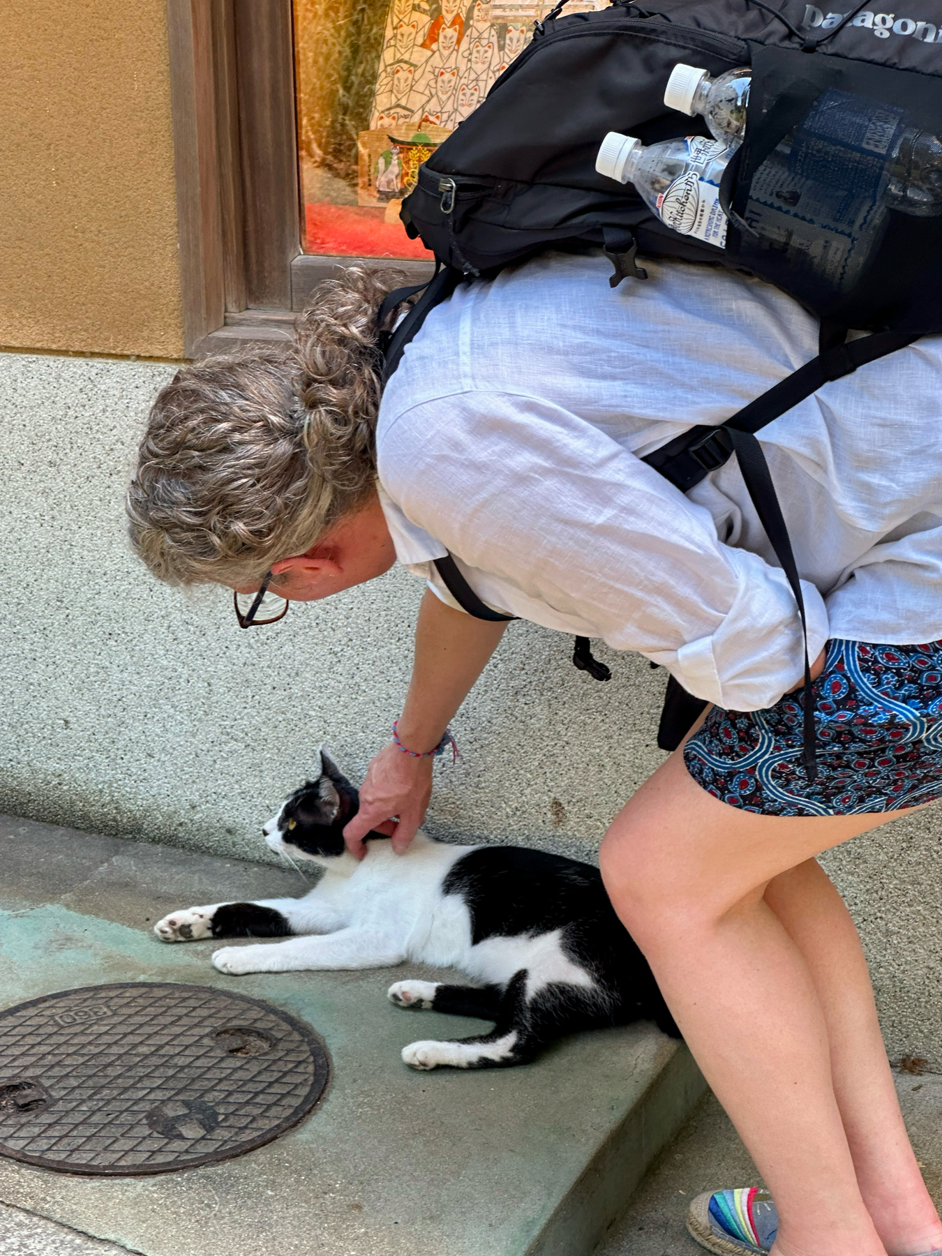 My wife crouched down petting a black and white cat lying on a doorstep, with a backpack and water bottles visible.