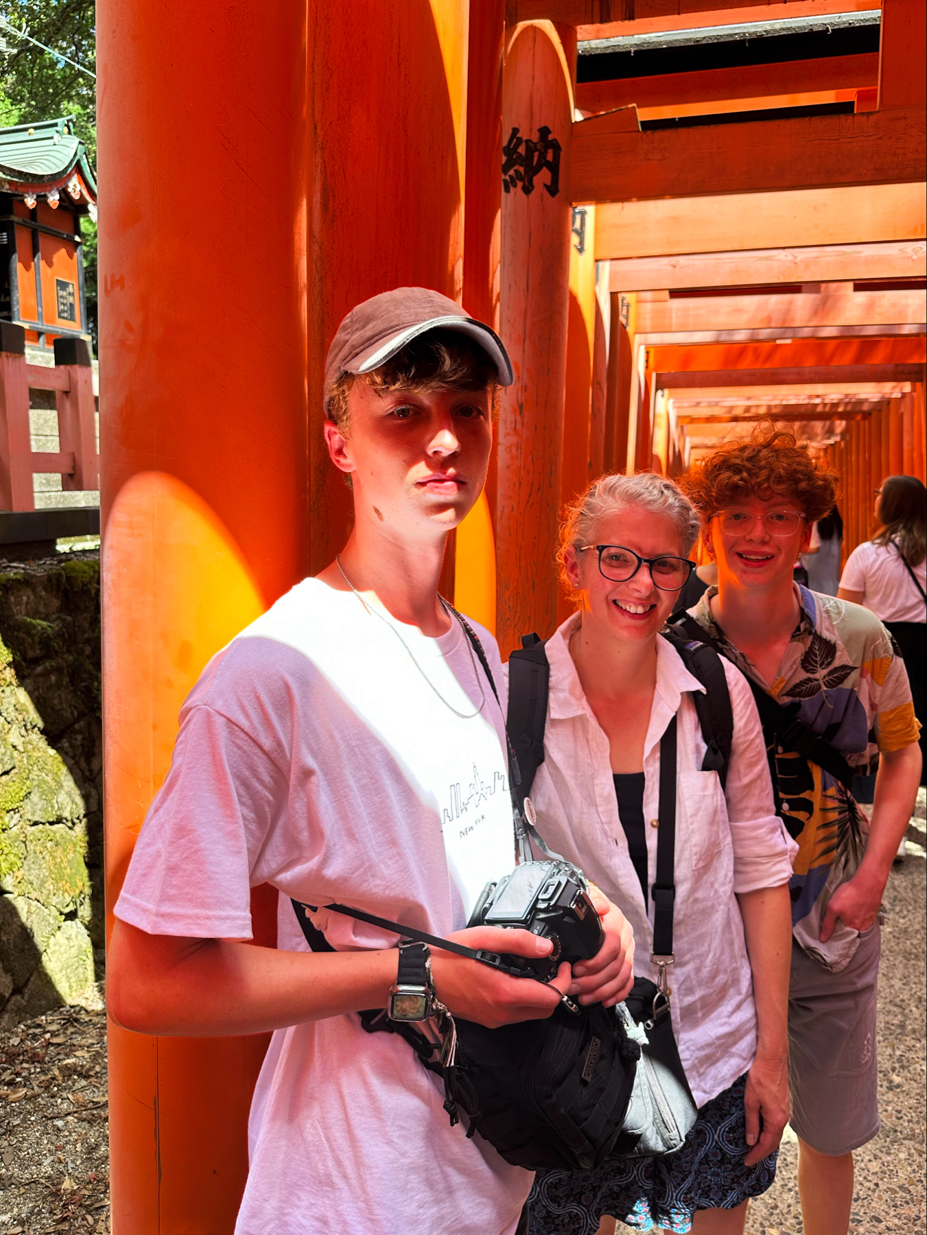My wife and two children stand among the vibrant orange torii gates at Fushimi Inari Shrine in Kyoto, Japan. The individual in the foreground is holding a camera, wearing a white T-shirt and cap, while the other two are smiling in the background,