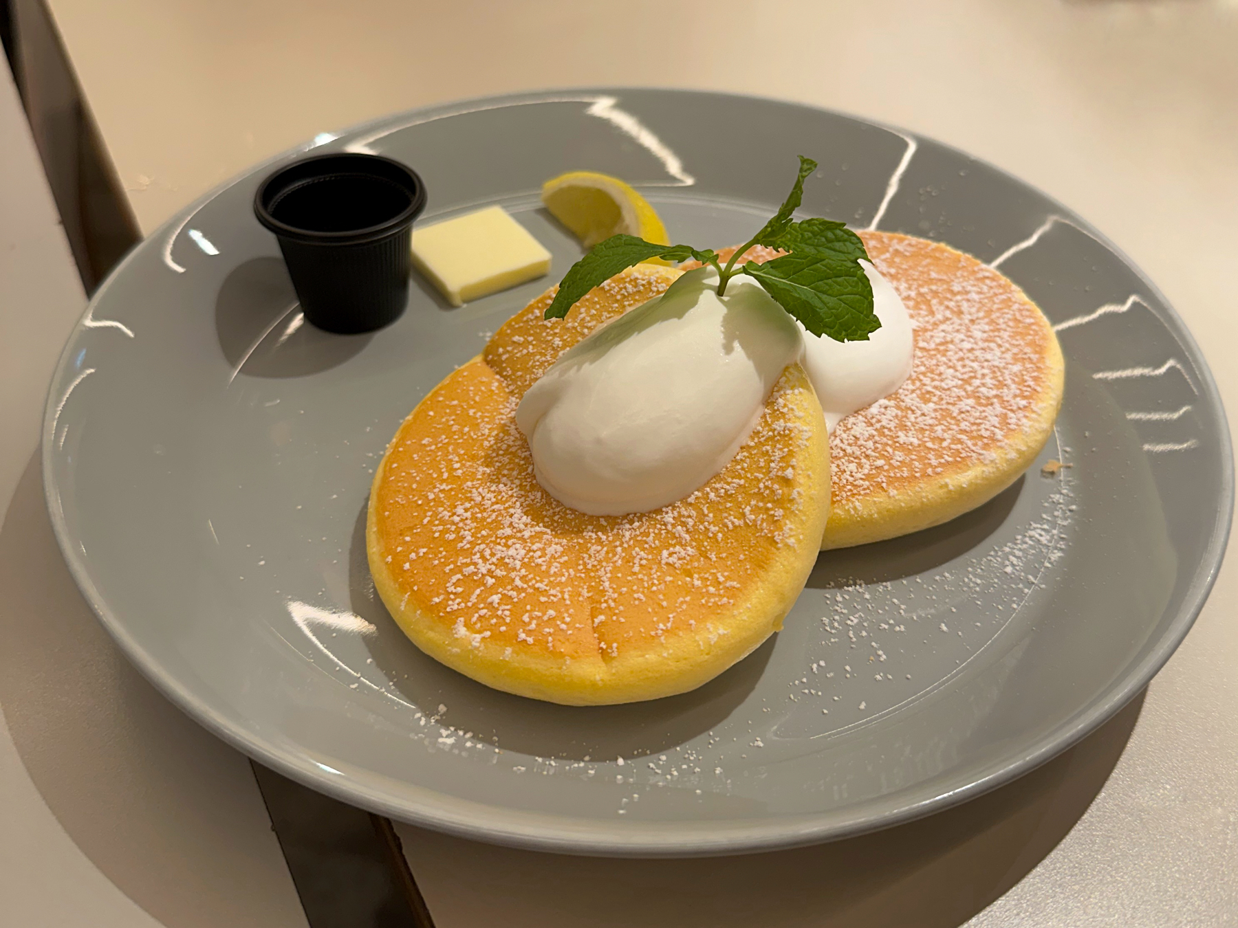 Two fluffy pancakes topped with a dollop of whipped cream, served with a slice of lemon, a pat of butter, and a small container of syrup, sprinkled with powdered sugar and garnished with a mint leaf, presented on a gray plate.